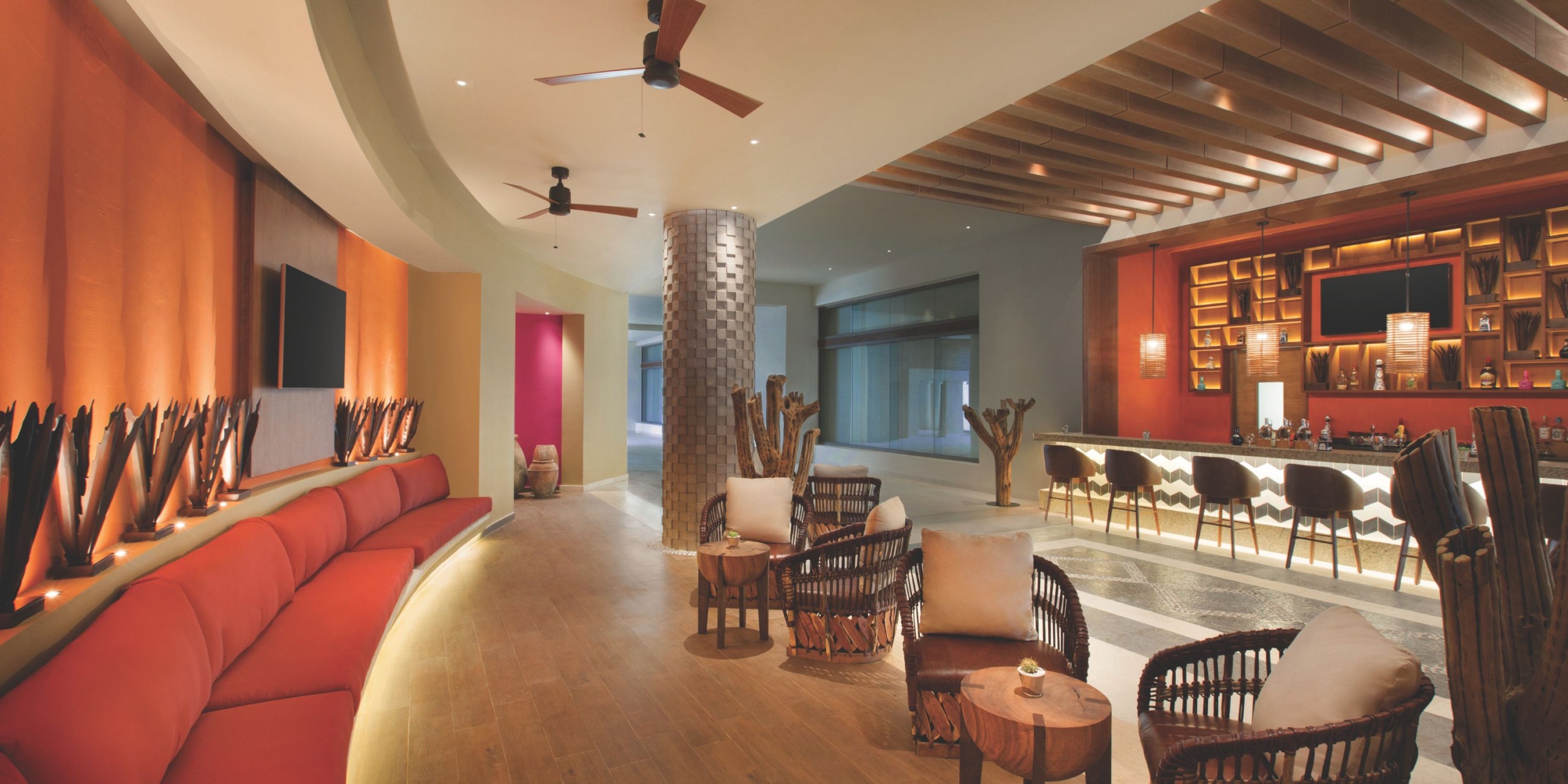 Hyatt Ziva Cancun features a modern lounge with red and tan seating, wicker chairs, a ceiling fan, and a bar adorned with wooden accents.