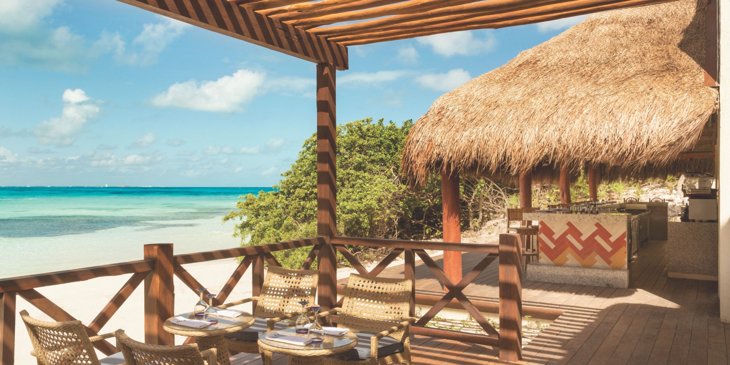 Hyatt Ziva Cancun offers a beachside patio with wooden furniture that overlooks the turquoise sea, all under a charming straw-roofed pergola. Nearby, guests can enjoy a tiki bar that is partly shaded by trees.
