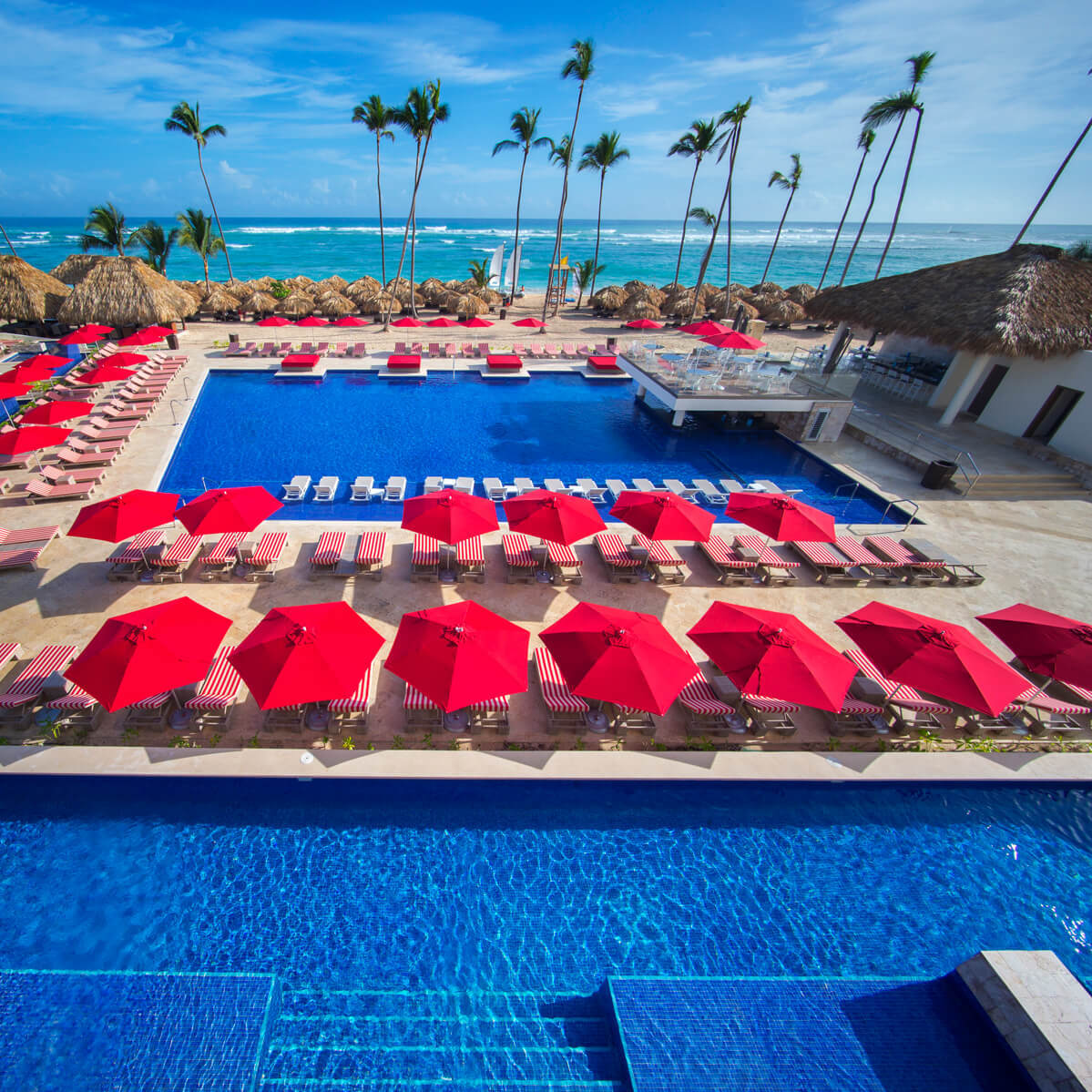A Royalton Bavaro, perfect for destination weddings, with a large blue swimming pool surrounded by red umbrellas and palm trees, overlooking the ocean.