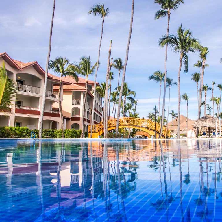 Majestic Colonial resort destination for weddings with tall palm trees, reflective blue swimming pool, and multi-story buildings under a clear blue sky.