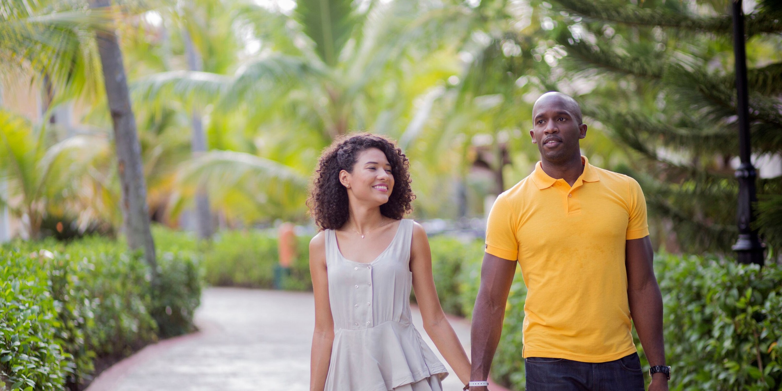 The couple strolls hand in hand along a tropical path at Majestic Elegance Punta Cana, surrounded by palm trees and lush greenery.