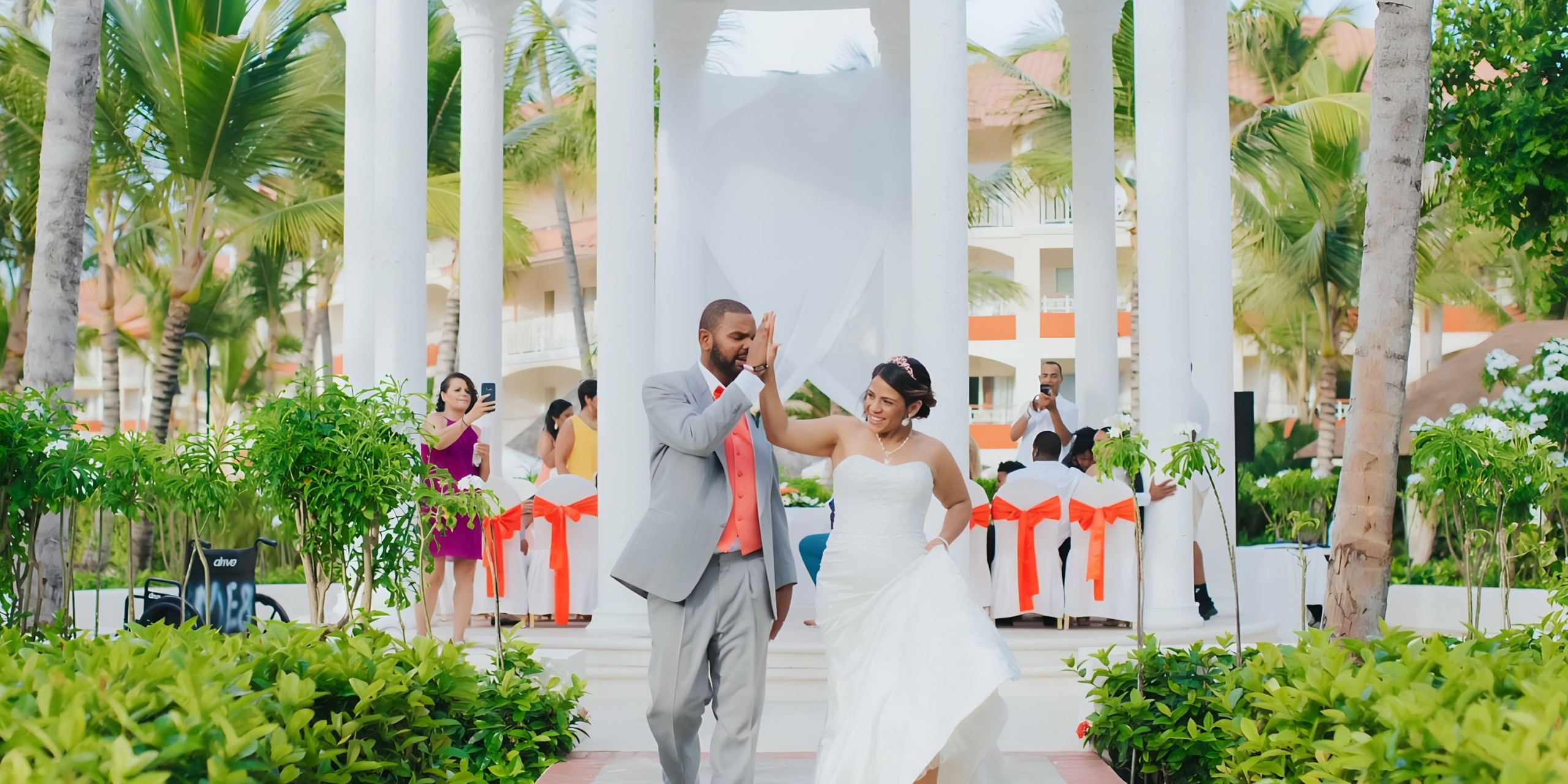 A newlywed couple dances amidst palm trees and columns at Majestic Elegance Punta Cana, with guests looking on.