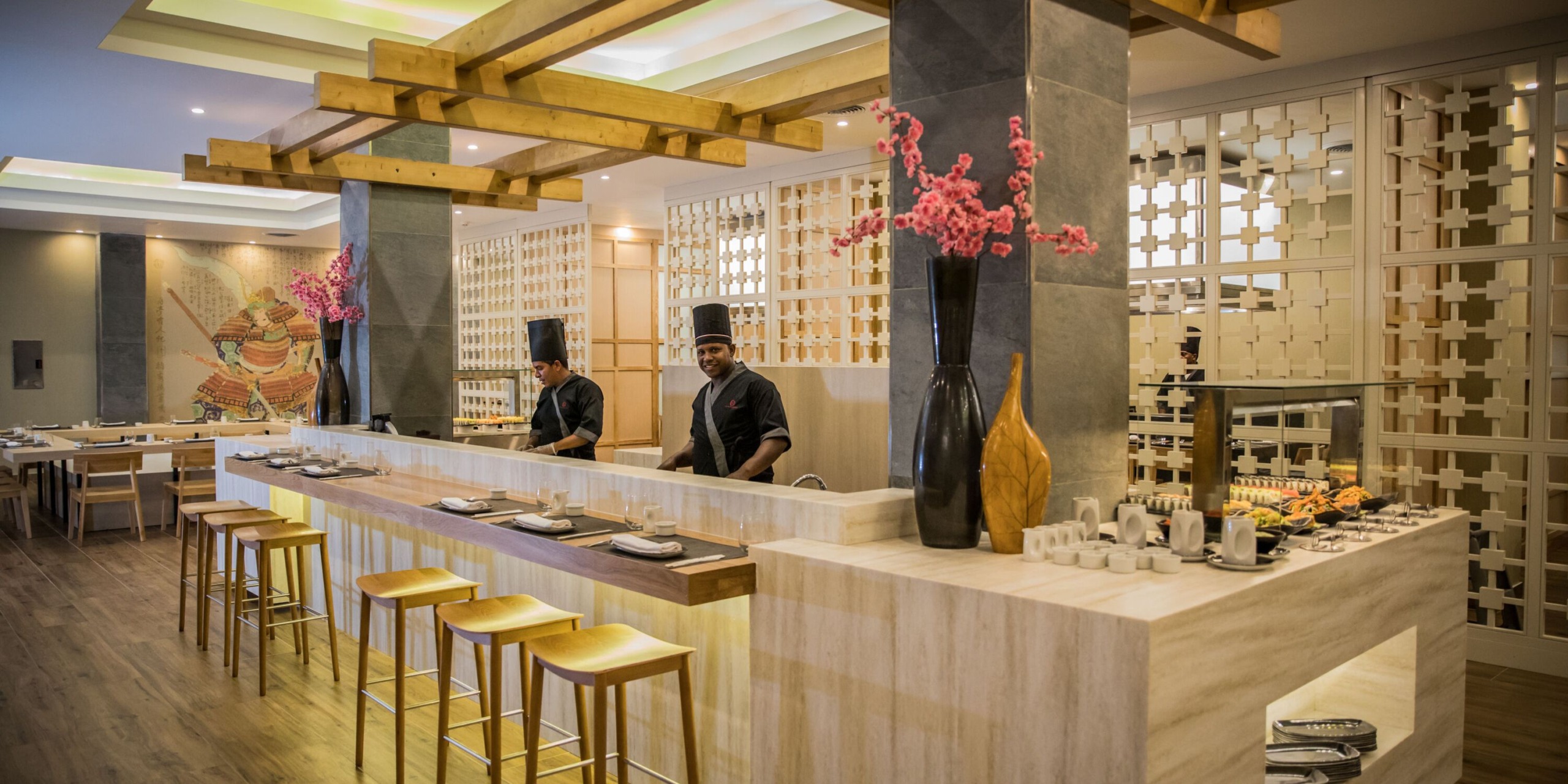 A sophisticated interior at Majestic Elegance Punta Cana features a sushi bar and wooden stools, with two chefs stationed behind the counter and pink flowers adding decorative flair to the ambiance.