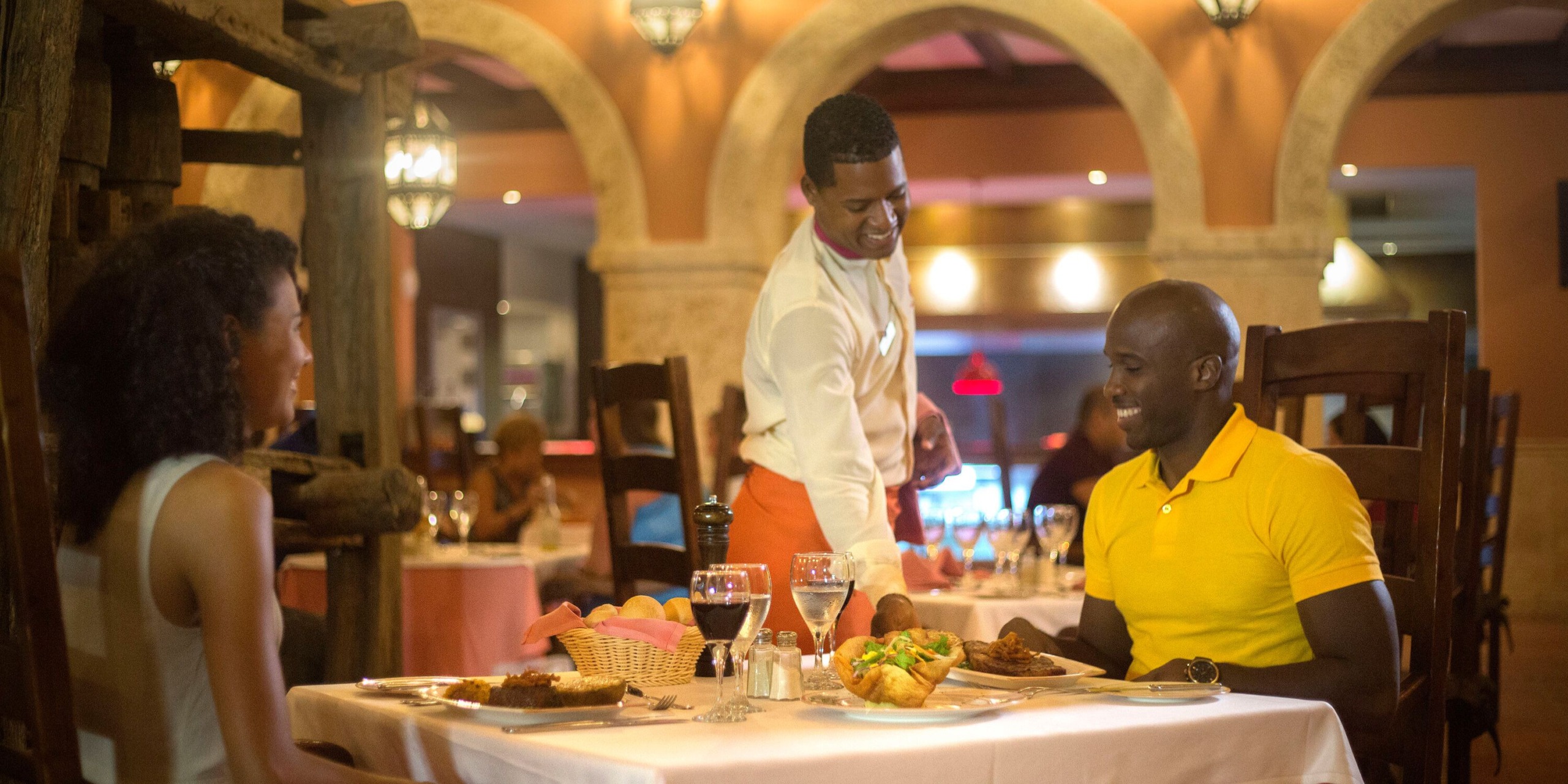 A waiter serves food to a man in a yellow shirt at a restaurant table in the Majestic Elegance Punta Cana, while a woman sits across from him.