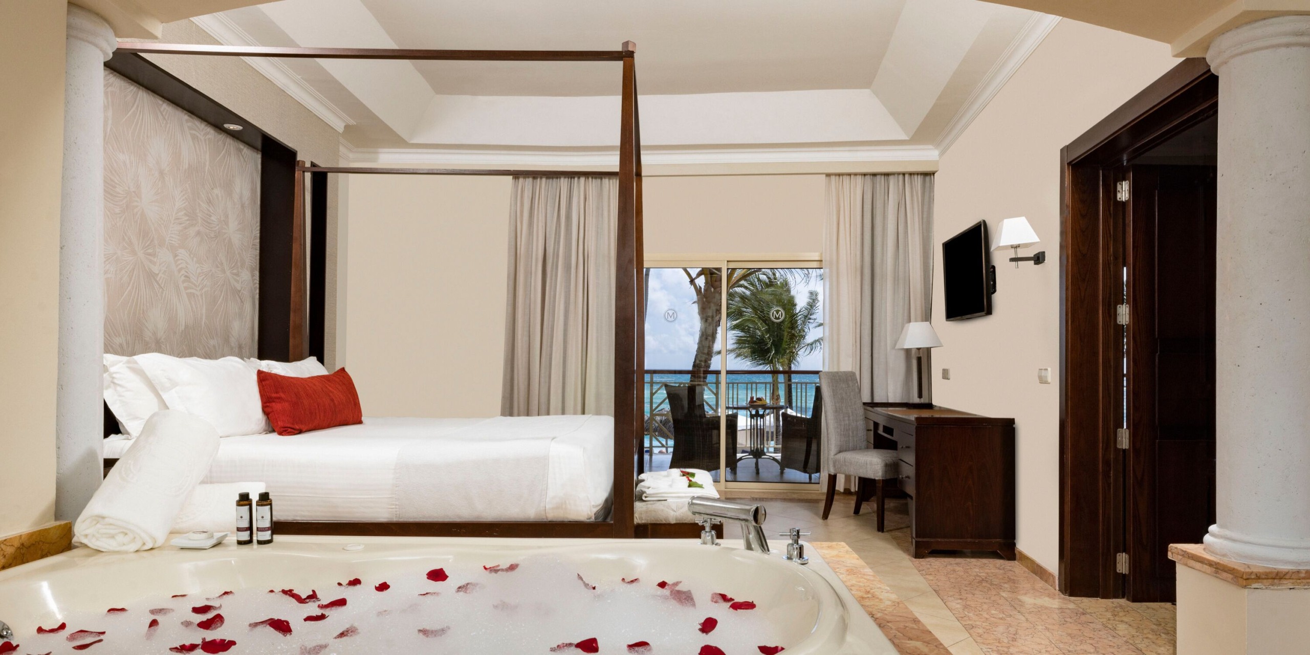Stay in the Majestic Elegance Punta Cana, featuring a luxurious four-poster bed and a jacuzzi tub sprinkled with rose petals. Enjoy breathtaking ocean views from your private balcony. The room is equipped with a desk, chair, and wall-mounted TV, all harmoniously set against a neutral color scheme enhanced by elegant red accents.