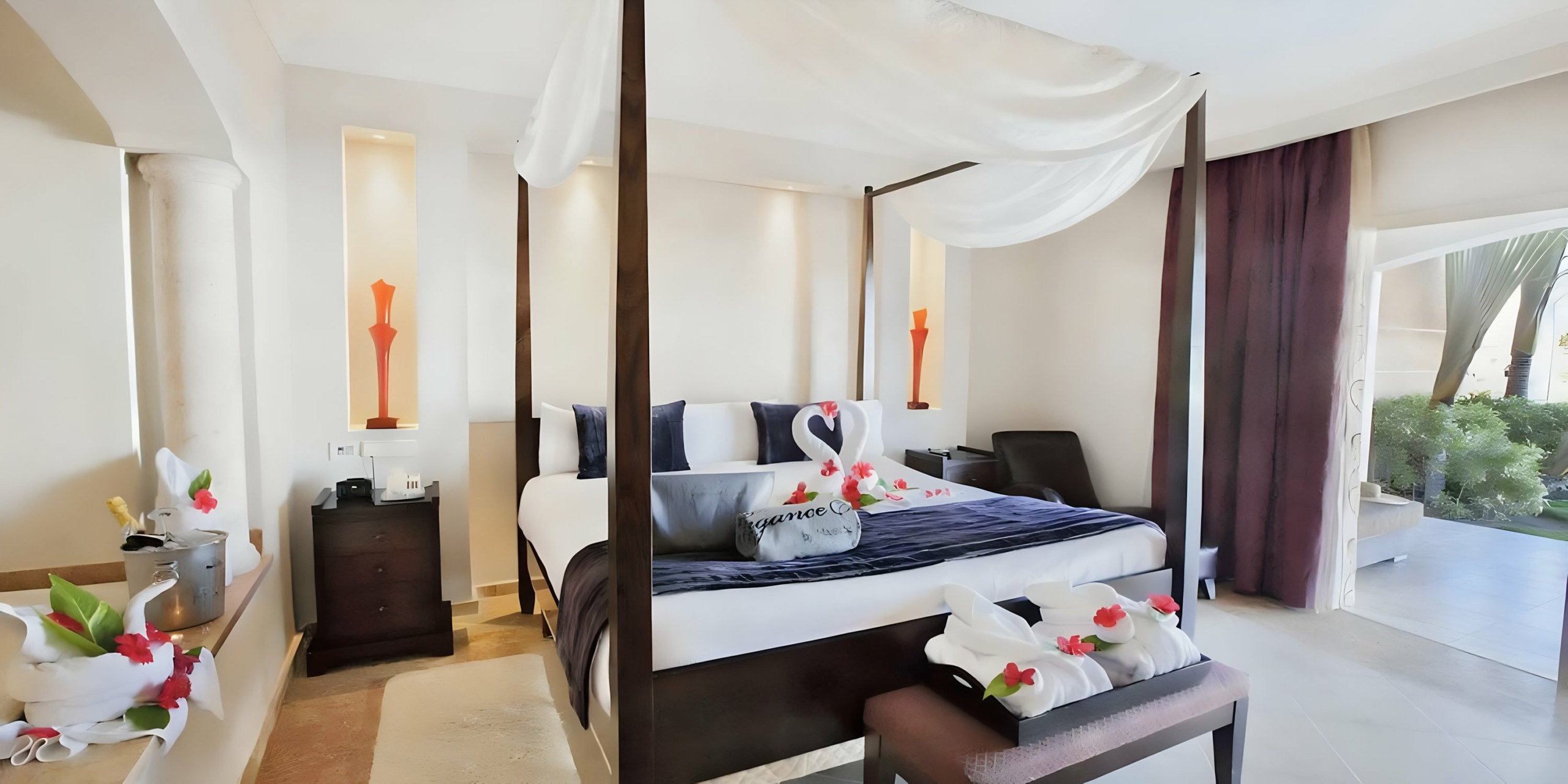 A modern bedroom with a four-poster bed, decorated elegantly with a white canopy, flower petals, and a heart-shaped arrangement of towels at the Majestic Elegance Punta Cana.