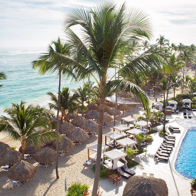 Majestic Elegance Punta Cana ideal for destination weddings, with thatched umbrellas, palm trees, sun loungers by a pool, and a view of the ocean.