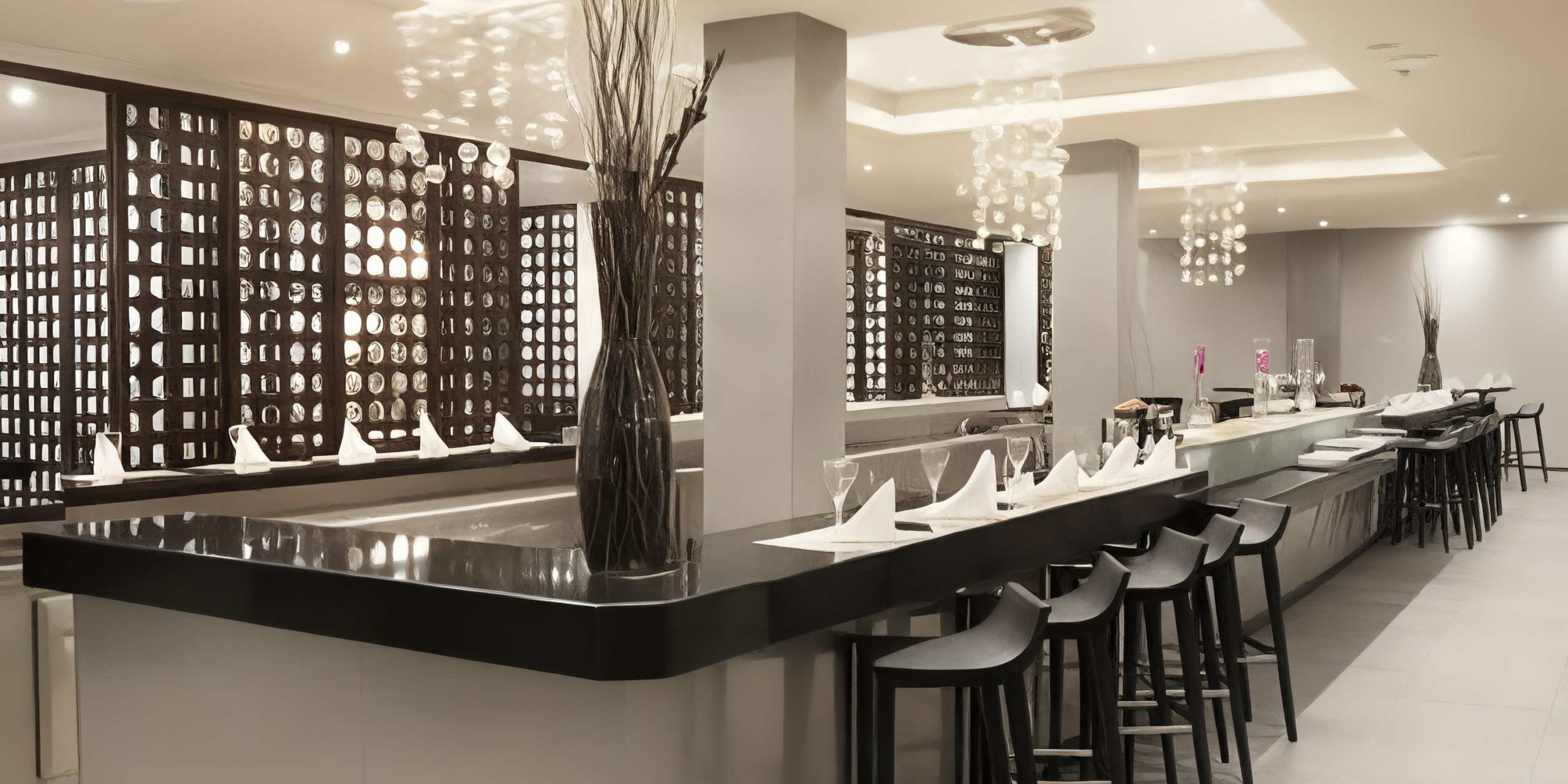 Majestic Elegance Punta Cana features a contemporary interior with a spacious bar counter, high stools, and sophisticated table arrangements. Neutral tones, artistic partitions, and overhead lighting contribute to the chic ambiance.