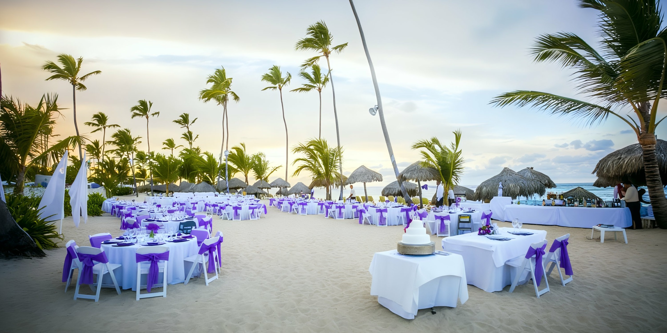 At the Majestic Elegance Punta Cana, an outdoor beach wedding setup features white tables and chairs adorned with purple ribbons, all surrounded by palm trees against a sunset sky. In the foreground, a tiered cake sits elegantly on a table.
