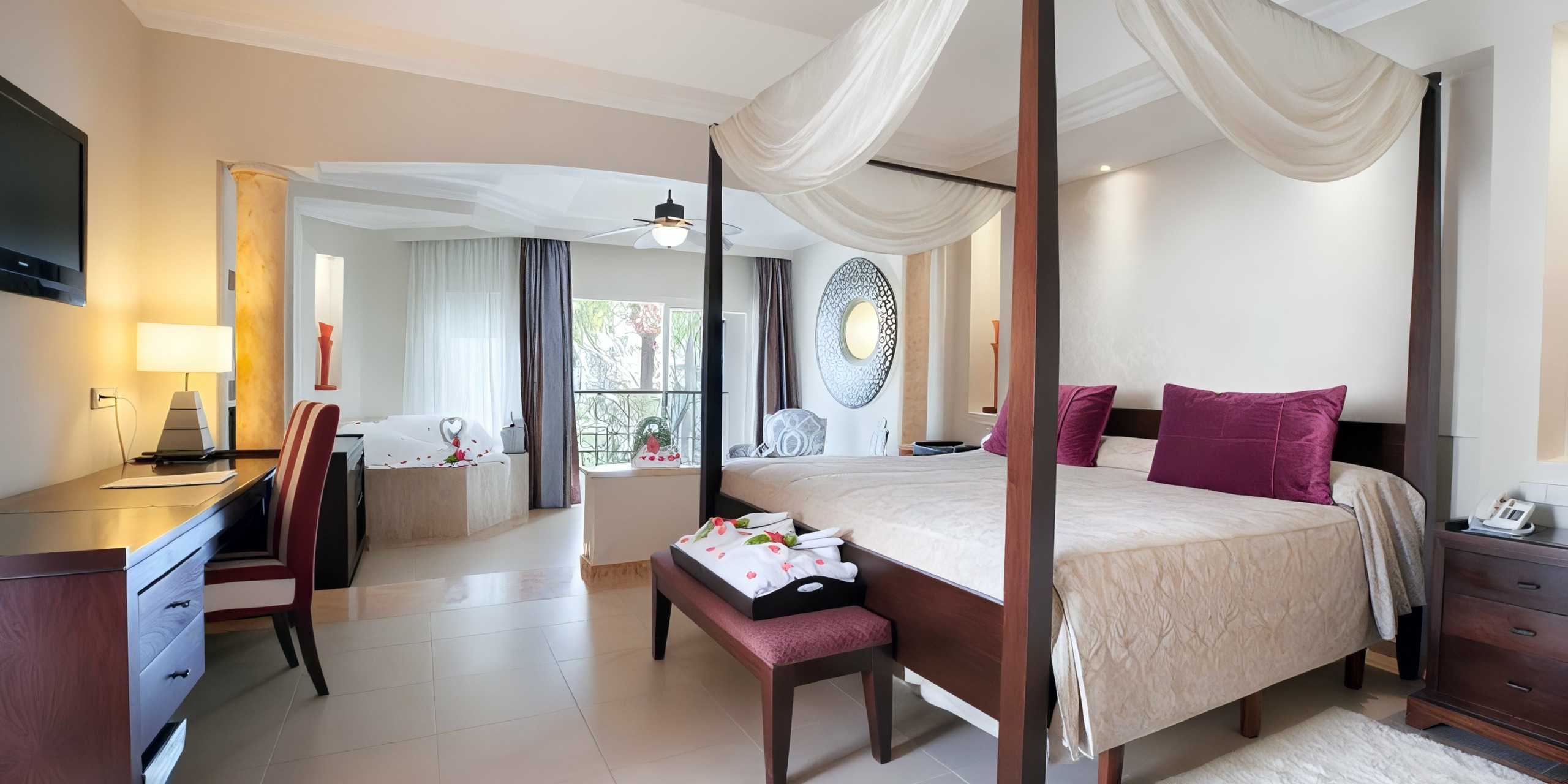 The Majestic Elegance Punta Cana offers a spacious hotel room featuring a four-poster bed, red cushions, a desk, and a balcony with stunning garden views. A jacuzzi is conveniently situated in the background near the balcony.