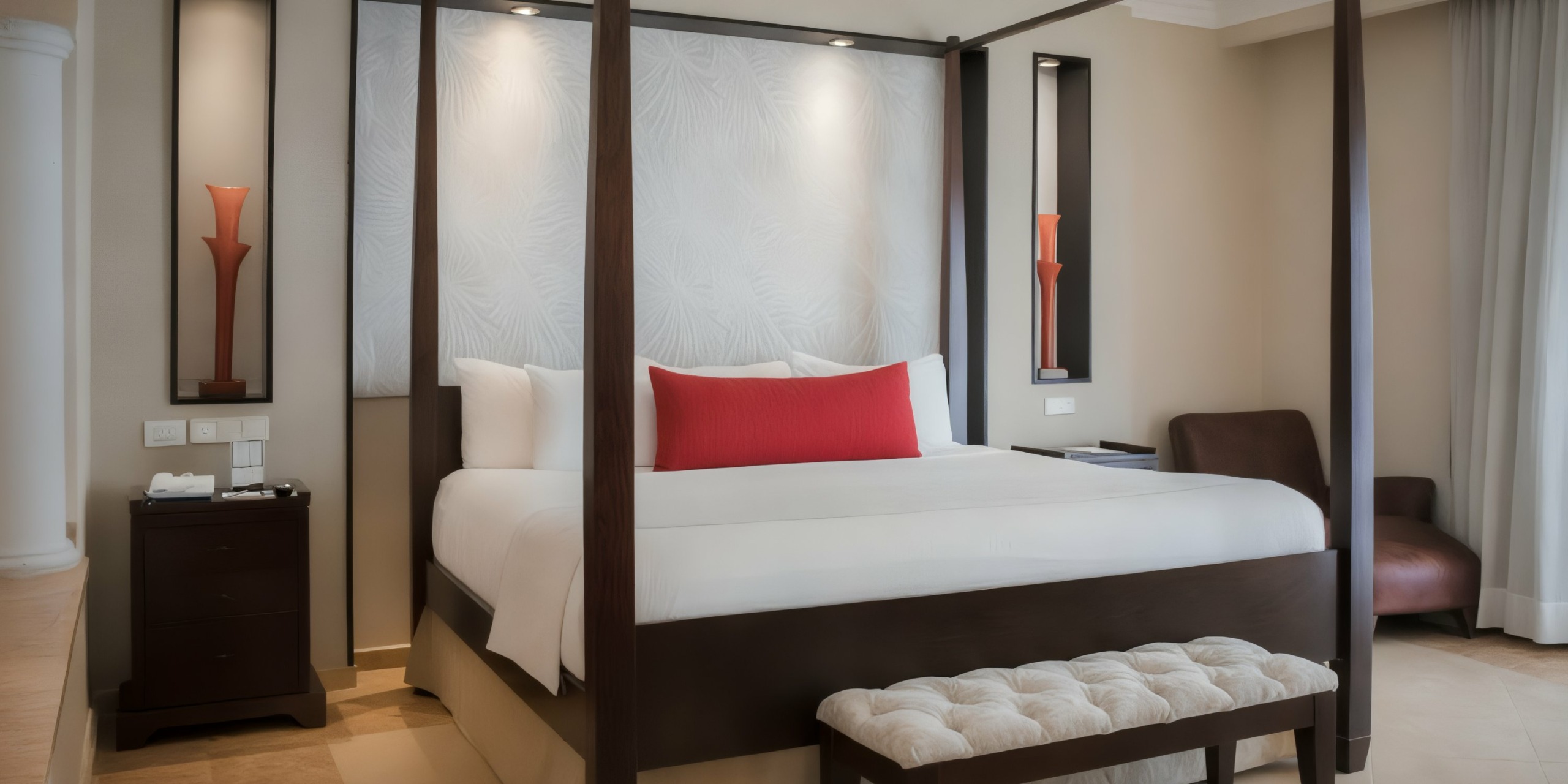 The Majestic Elegance Punta Cana hotel room features a four-poster bed with a red accent pillow, a bedside table, and a bench at the foot of the bed. A leather chair occupies the corner, complemented by decorative wall panels.