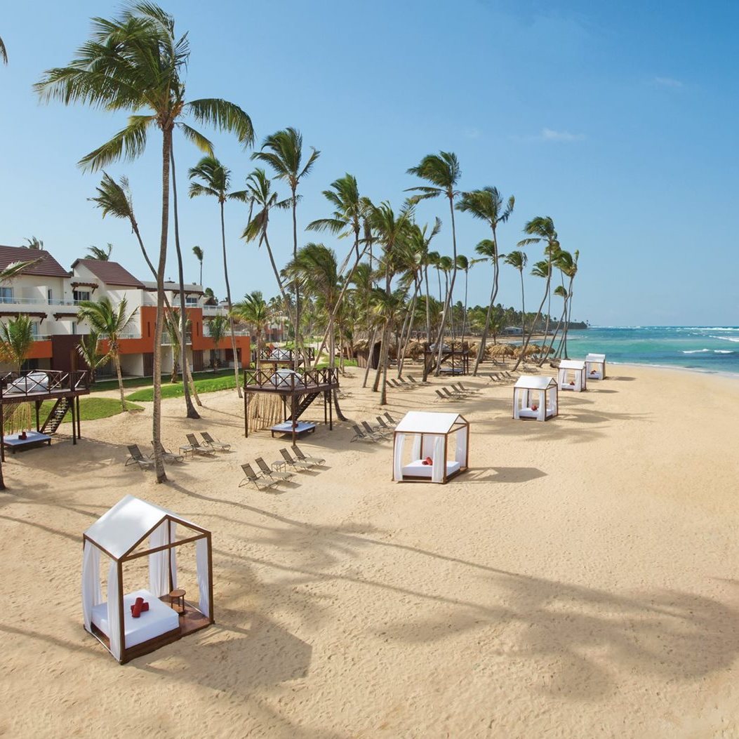 A tropical beach with palm trees, cabanas, lounge chairs, and Breathless Punta Cana Resort & Spa ideal for destination weddings in the background under a clear blue sky.