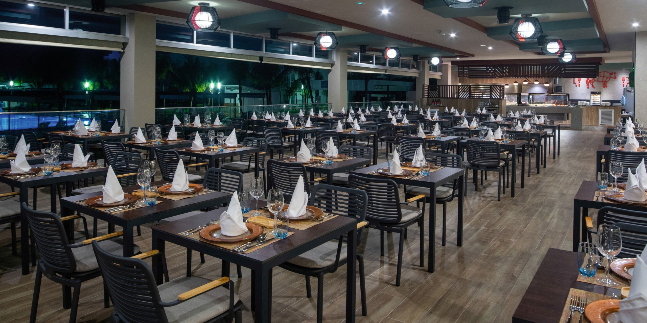 The Hotel Riu Montego Bay boasts a spacious dining area with rows of neatly arranged tables, each elegantly adorned with white napkins and glassware. The ceiling is decorated with lantern-style lighting, while large windows provide a scenic outdoor view.