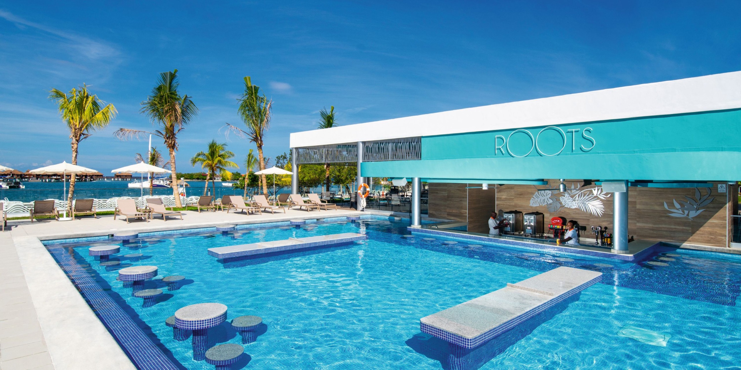 The outdoor pool area at Hotel Riu Montego Bay features a swim-up bar named "ROOTS," with sun loungers and palm trees set against a backdrop of a clear blue sky.