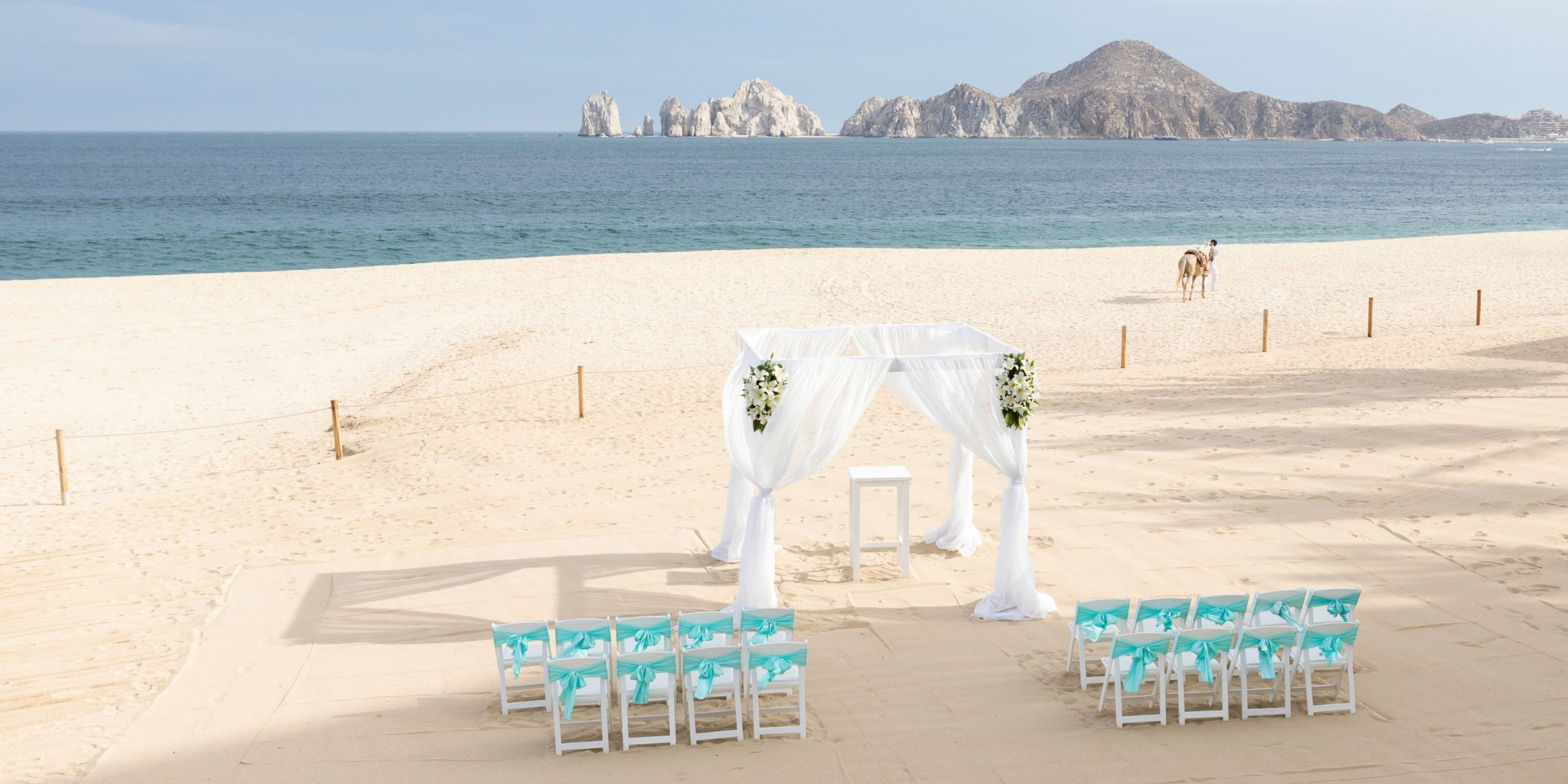 A white wedding canopy and chairs are elegantly set up on the sandy beach of Hotel Riu Palace Cabo San Lucas, offering a picturesque distant view of the ocean and mountains.