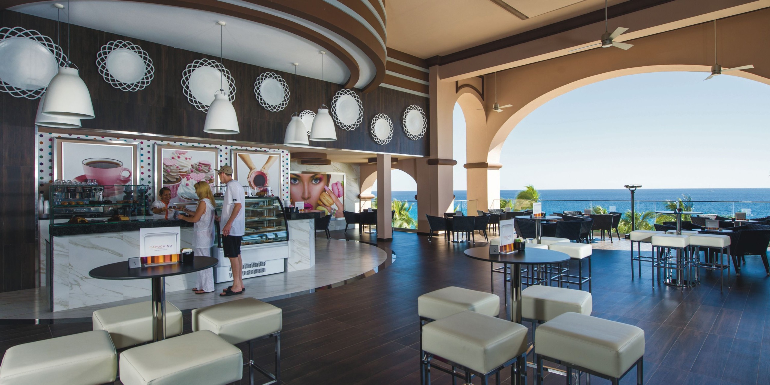 The interior of Hotel Riu Palace Cabo San Lucas showcases a contemporary cafe design where guests gather at the counter. The area is characterized by high ceilings and pendant lighting, complemented by expansive views of the ocean framed by large arches. Tables and chairs are thoughtfully placed on an elegant dark wood floor.