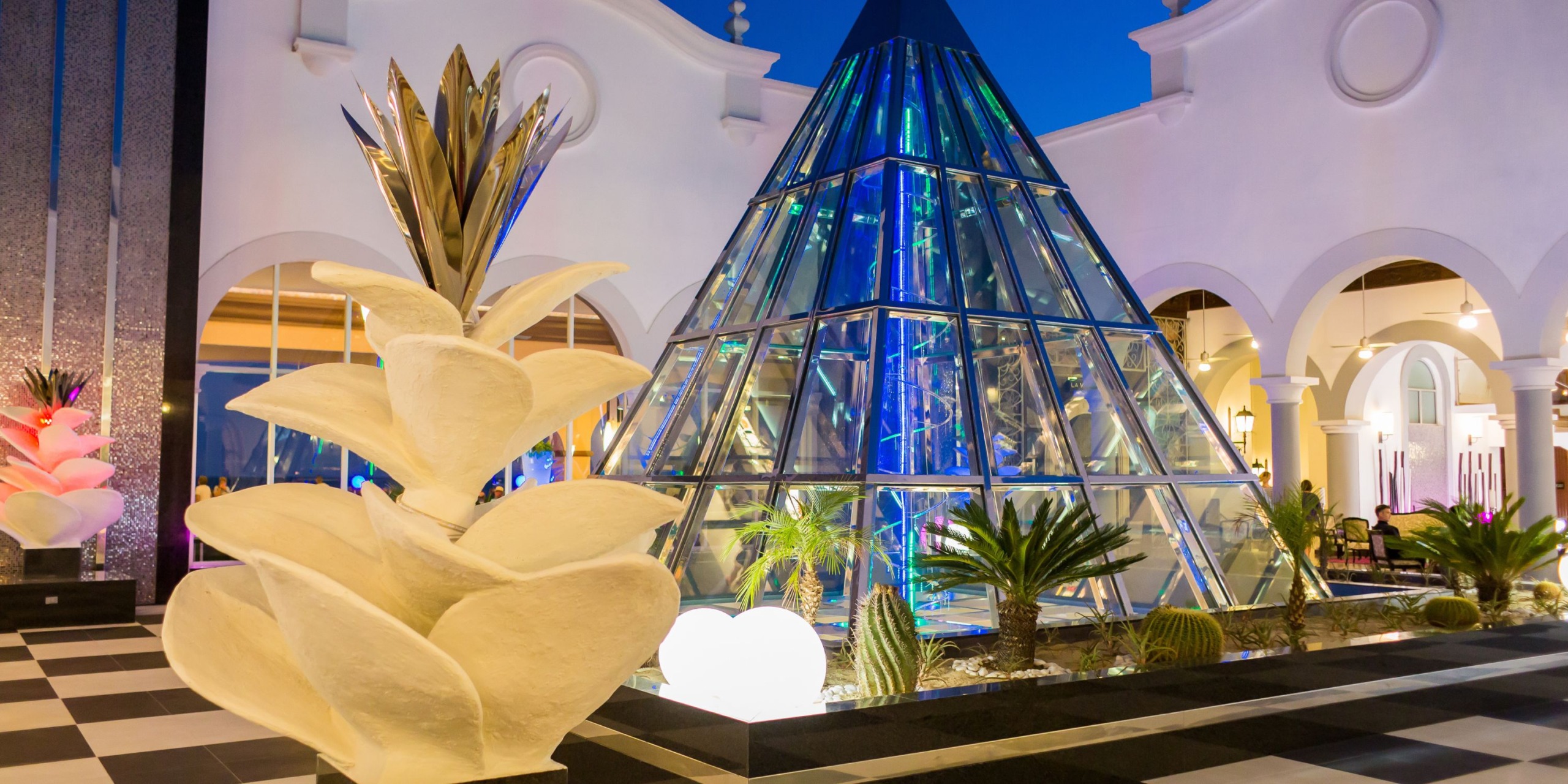 The Hotel Riu Palace Cabo San Lucas features a contemporary glass pyramid structure illuminated with blue lights, set among arches and a large abstract sculpture in its outdoor plaza.