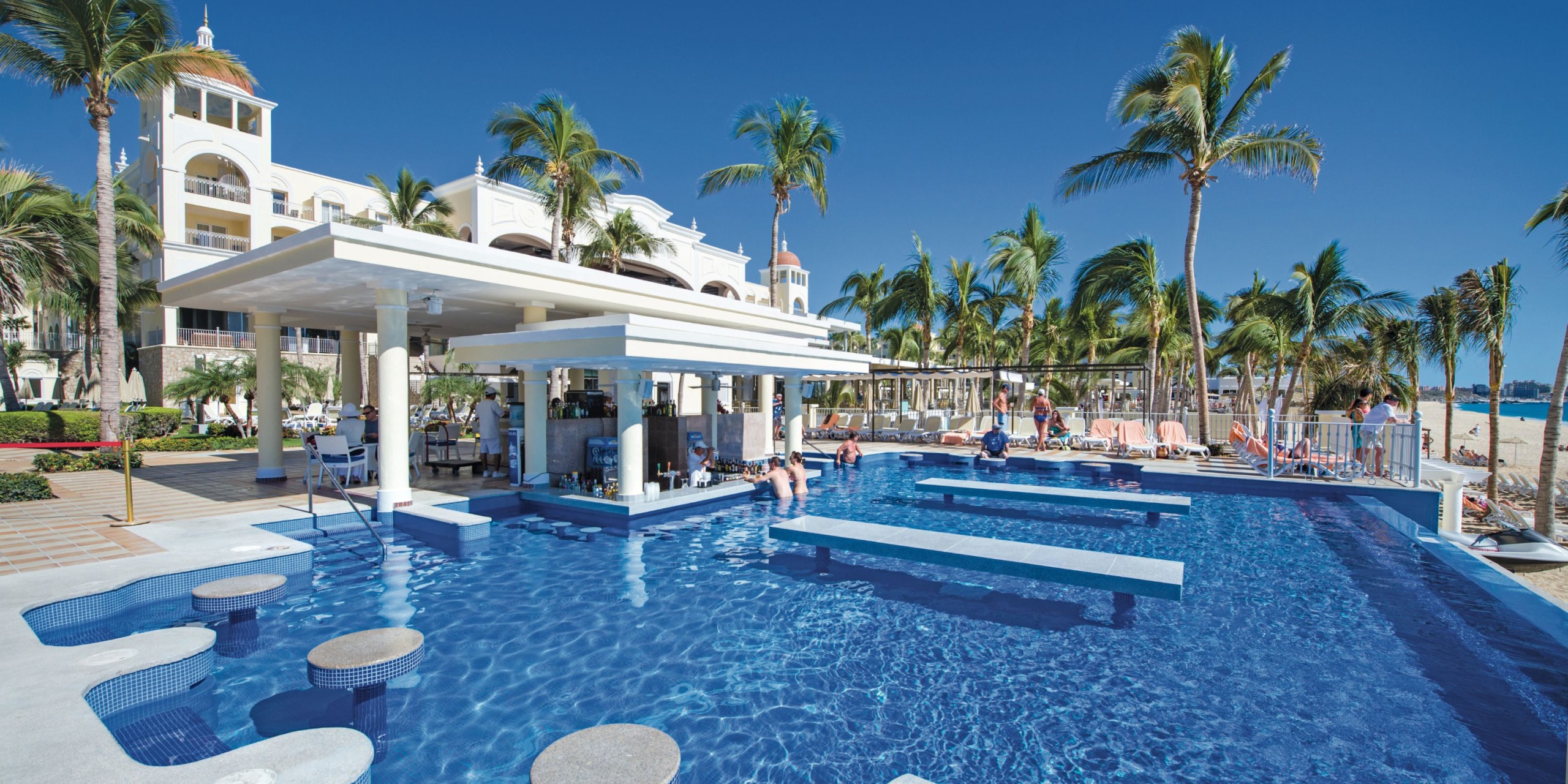 The Hotel Riu Palace Cabo San Lucas features an outdoor pool area with a swim-up bar, surrounded by palm trees. Guests are enjoying relaxation in and around the pool under a clear blue sky.