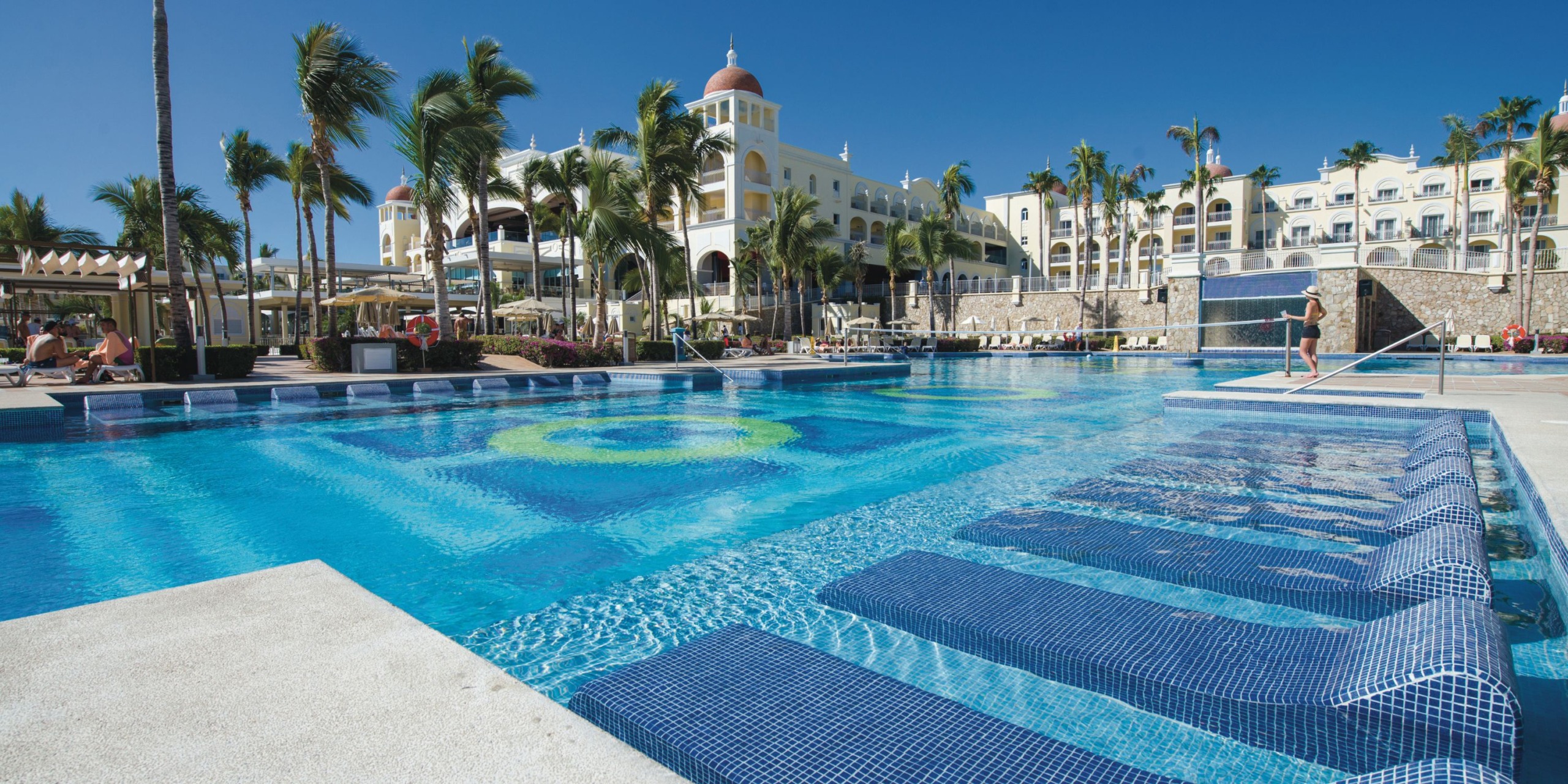 The Hotel Riu Palace Cabo San Lucas features an outdoor swimming pool equipped with submerged lounge chairs, surrounded by palm trees, with a large hotel building as the backdrop, all beneath a clear blue sky.