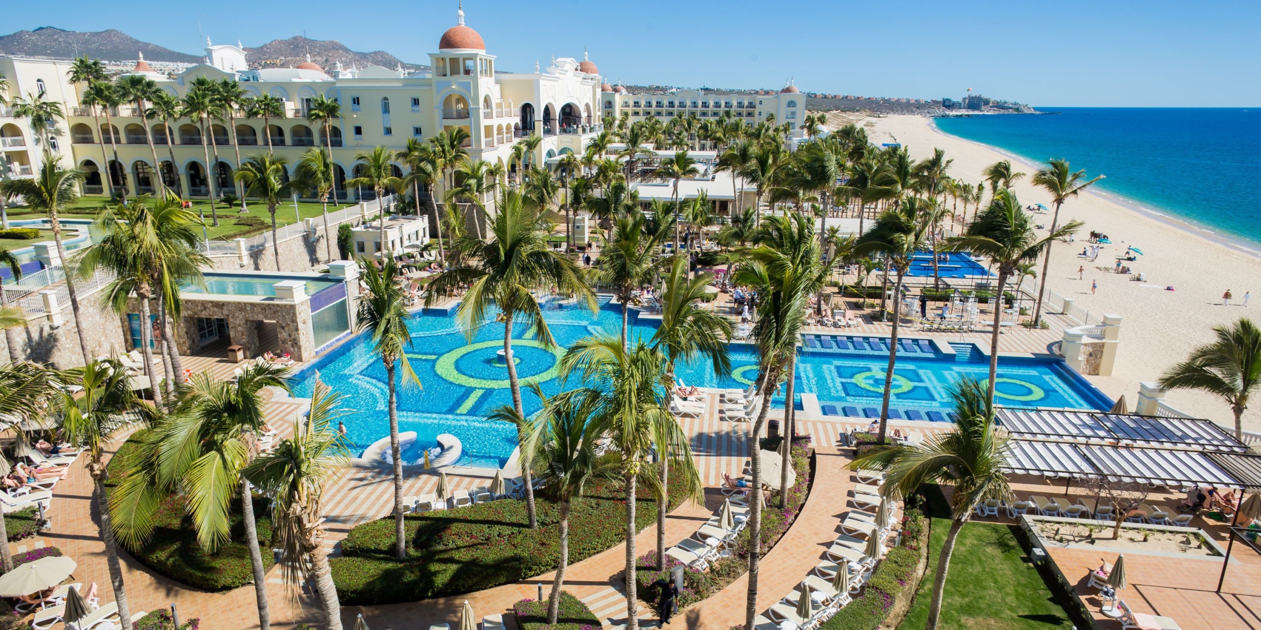Aerial view of the Hotel Riu Palace Cabo San Lucas, showcasing its expansive pool, palm trees, and inviting lounge areas. The sandy beach and ocean provide a stunning backdrop.
