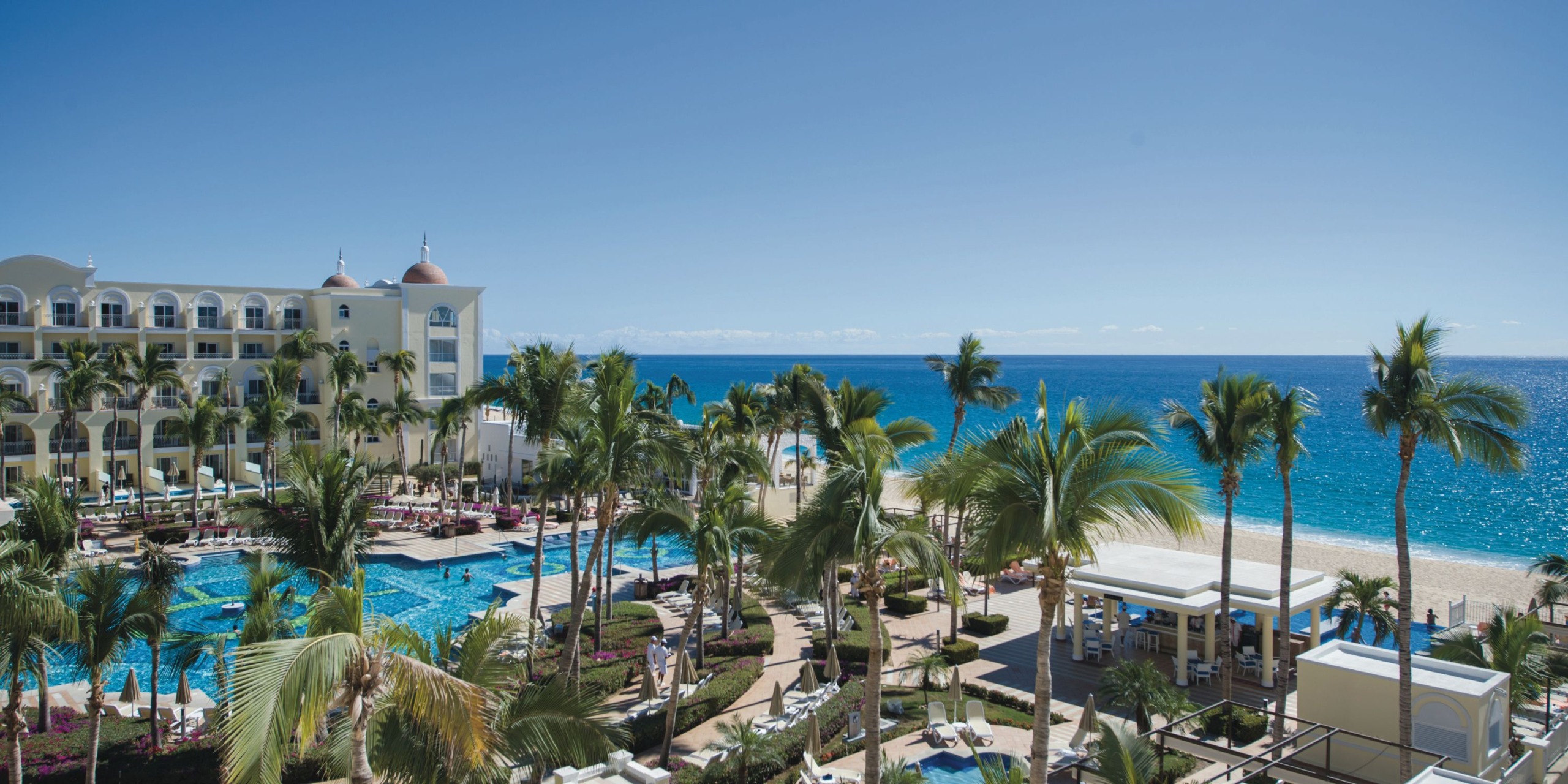 Experience the serene beauty of Hotel Riu Palace Cabo San Lucas, featuring palm trees, a spacious pool, comfortable lounge chairs, and the stunning backdrop of a bright blue ocean under a clear sky.