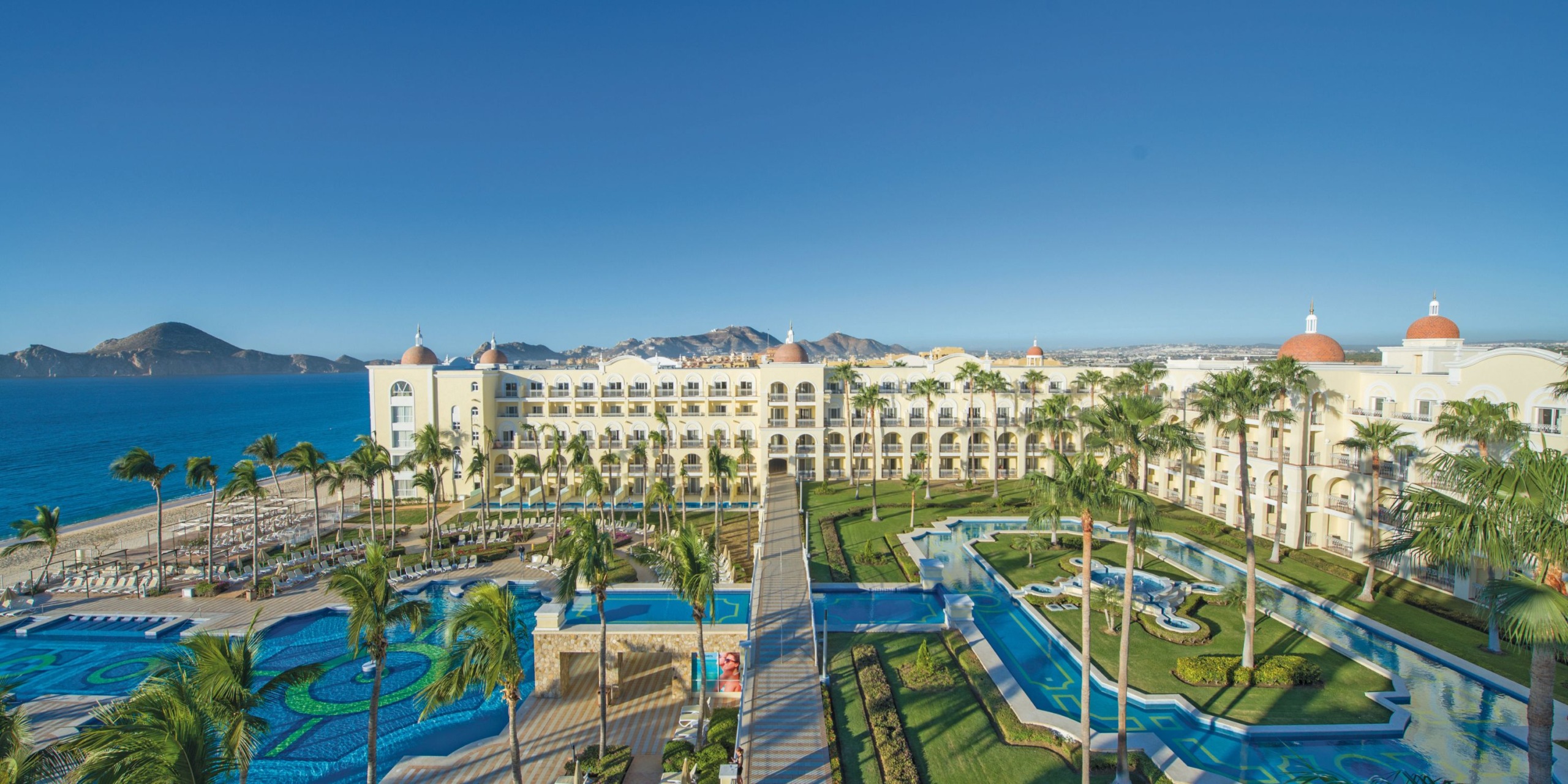 Aerial view of the Hotel Riu Palace Cabo San Lucas, a beachfront resort featuring a large hotel, multiple pools, palm trees, and the ocean in the background under a clear blue sky.