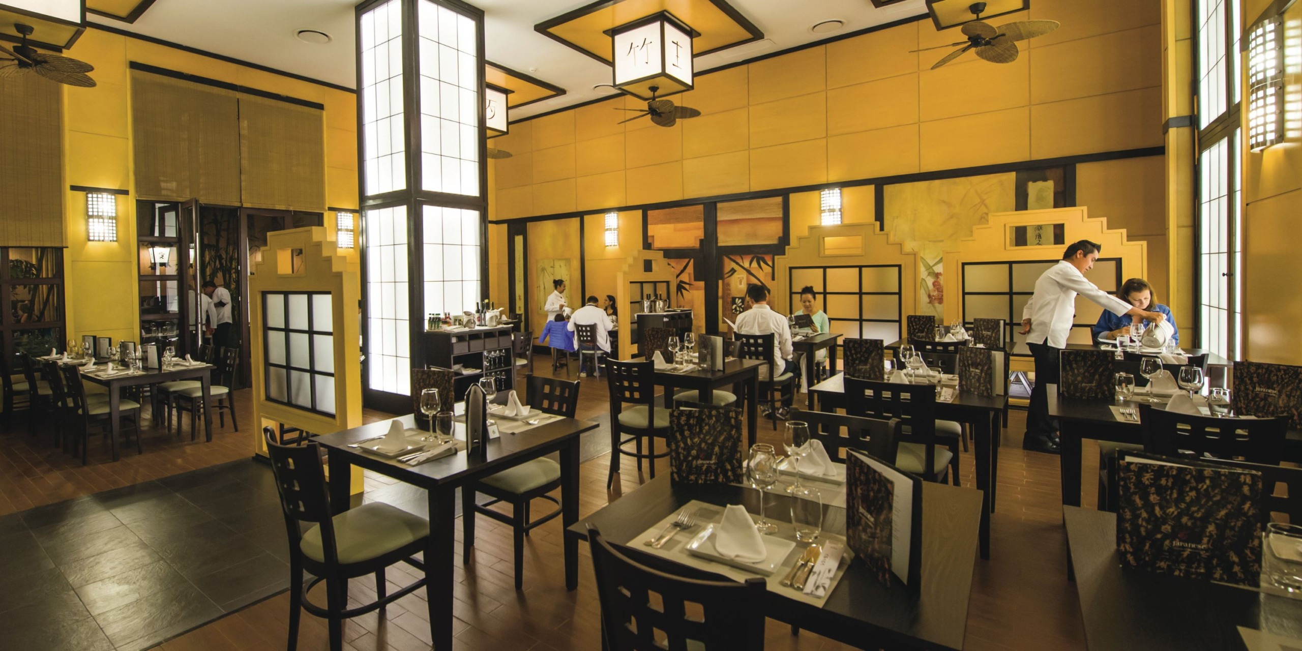 The interior of Hotel Riu Palace Cabo San Lucas boasts a spacious dining area with wooden furniture, set tables, and staff members diligently preparing for service, complemented by large windows and warm yellow walls.