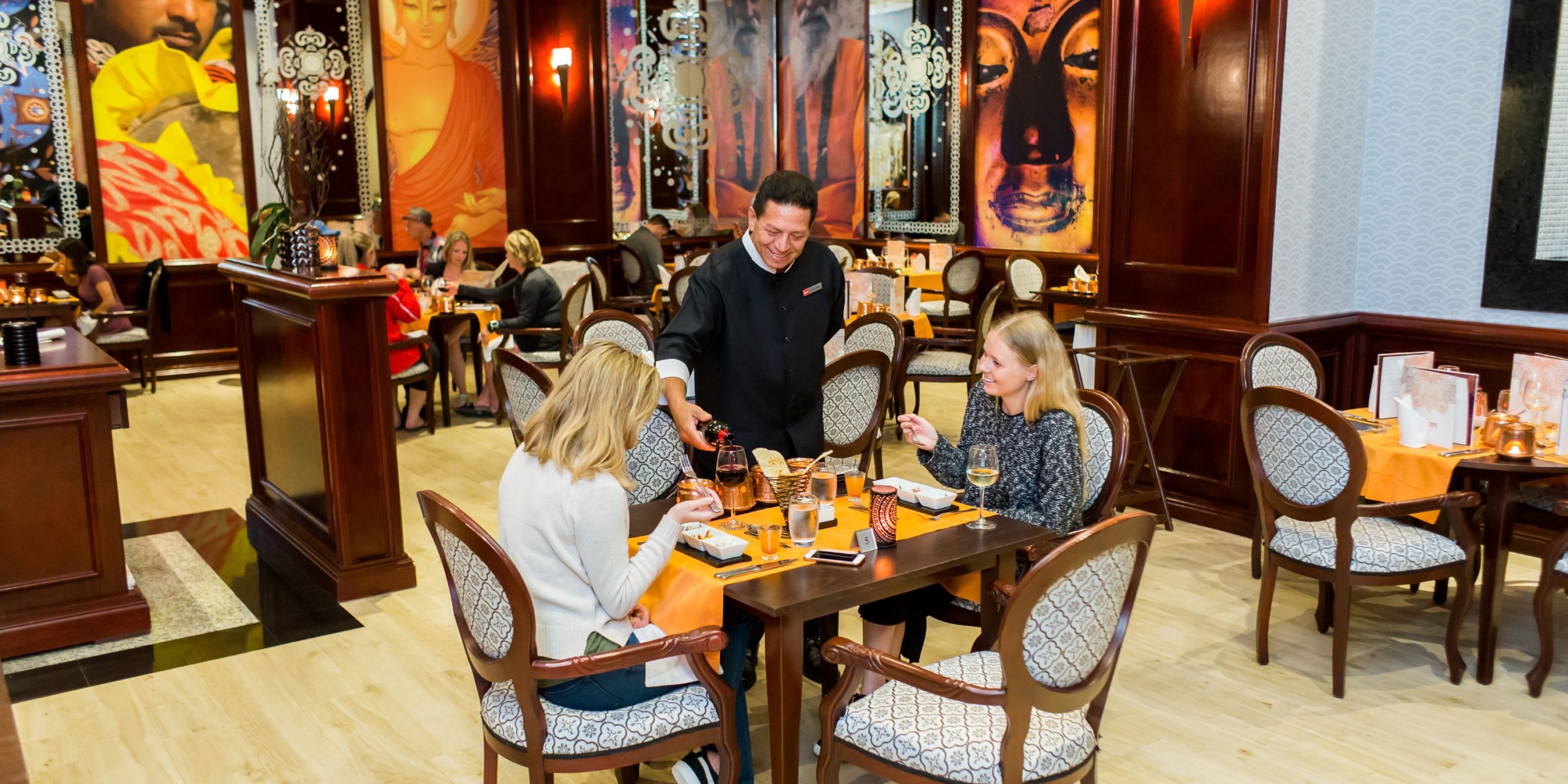 In the Hotel Riu Palace Cabo San Lucas, a waiter brings drinks to two women enjoying their meal amid the vibrant artwork on the walls.