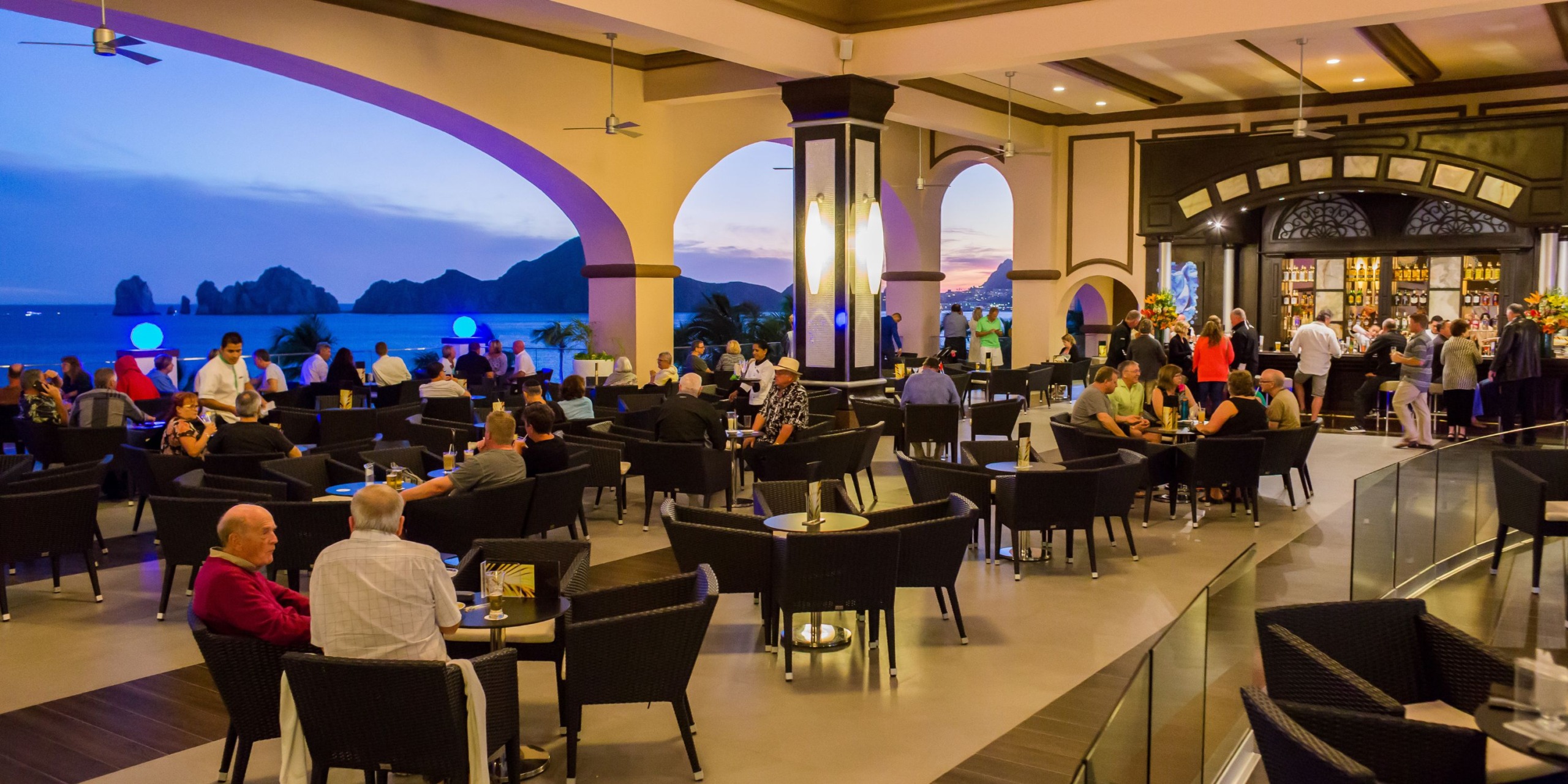 The lively indoor bar at Hotel Riu Palace Cabo San Lucas is filled with guests seated at tables, while large arched windows offer stunning views of the coastline at sunset.