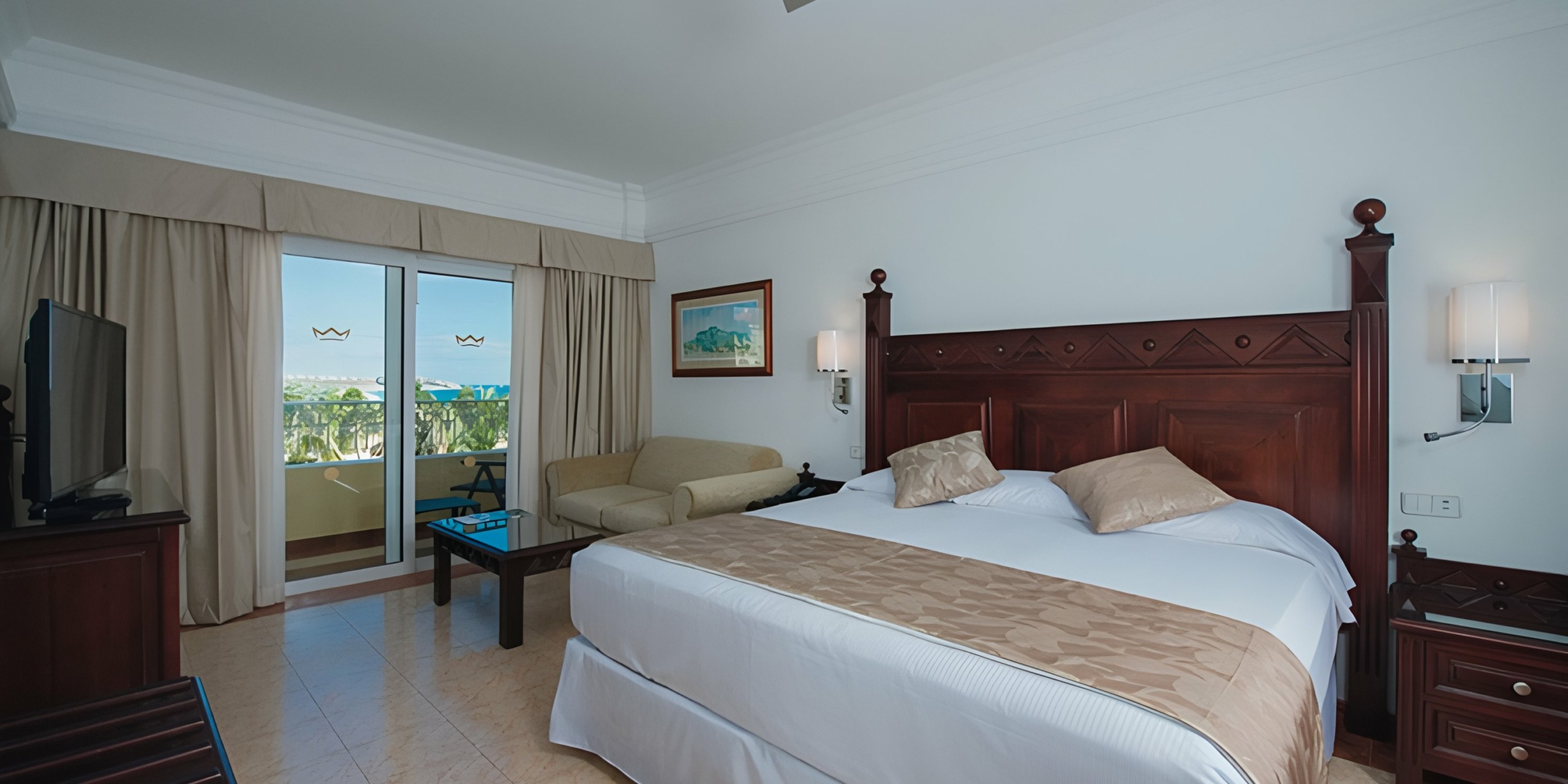 A spacious Hotel Riu Palace Cabo San Lucas room with a king-size bed, a seating area, and a balcony overlooking greenery.