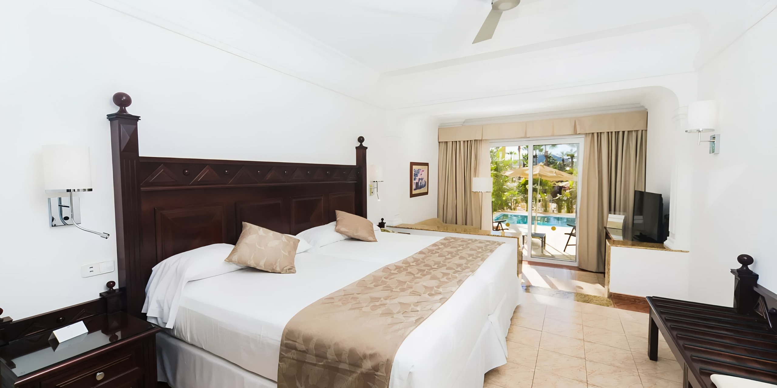 Bright and airy Hotel Riu Palace Cabo San Lucas room featuring a king-size bed, dark wood furniture, and sliding doors leading to a balcony with a pool view.