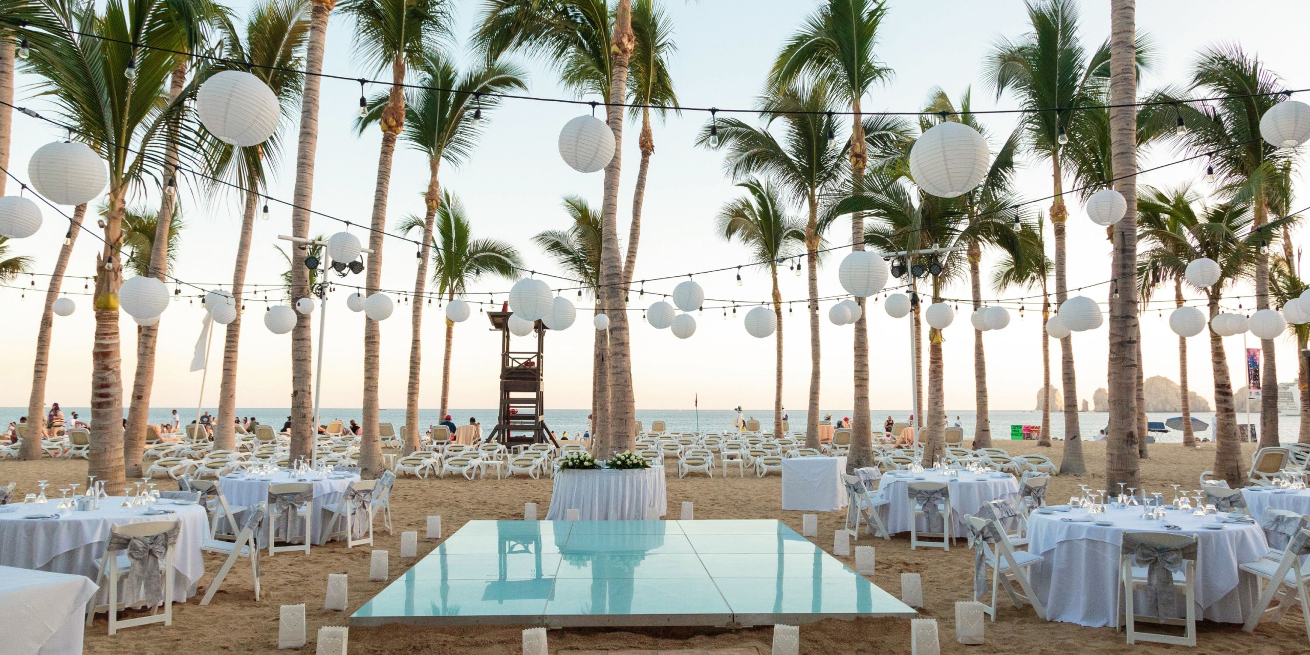 Hotel Riu Palace Cabo San Lucas offers a stunning beachside wedding setup featuring white tables and chairs, surrounded by palm trees and adorned with string lights and lanterns. A reflective platform is elegantly placed at the center, with the breathtaking ocean serving as a picturesque backdrop.