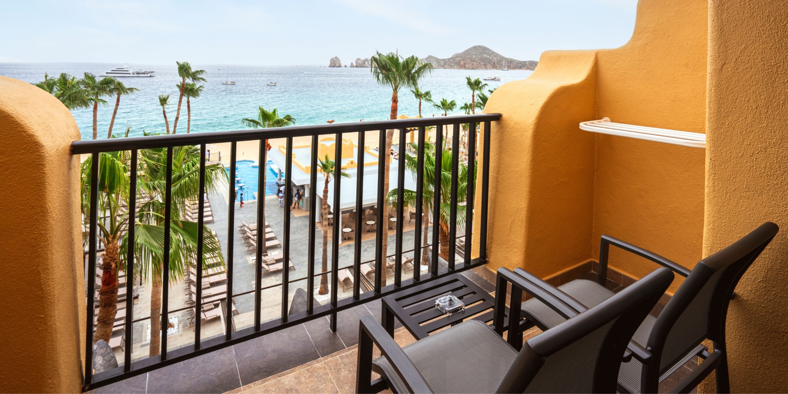 From the balcony of the Hotel Riu Santa Fe, you can enjoy a view overlooking a beach lined with palm trees. The balcony is furnished with two black chairs and a table. In the distance, boats gently glide across the water, framed by a rocky landscape.