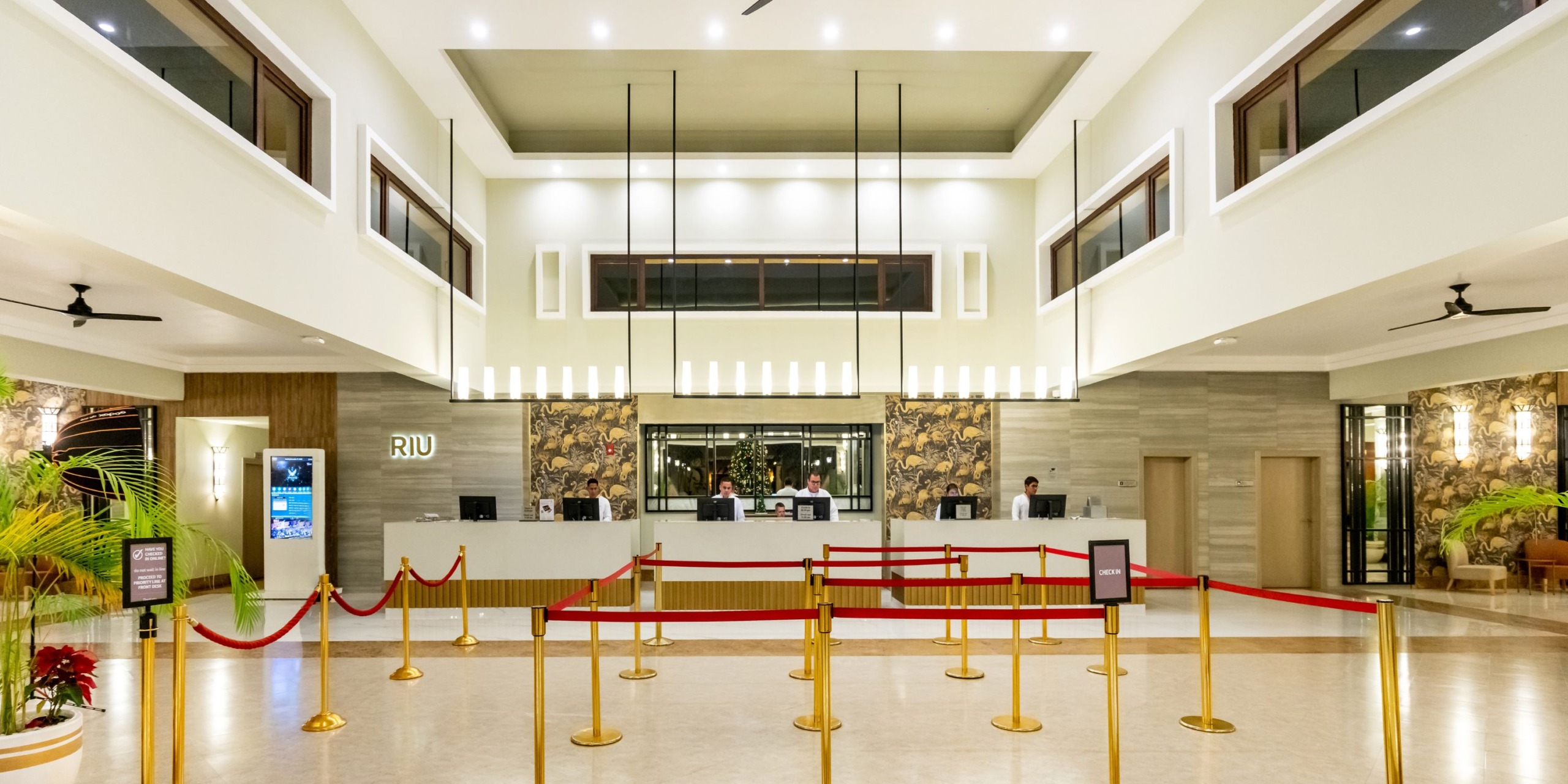 The Hotel Riu Santa Fe lobby features high ceilings, a modern design, and a front desk staffed by employees. Red ropes guide guests, and the Hotel Riu Santa Fe logo is prominently displayed.