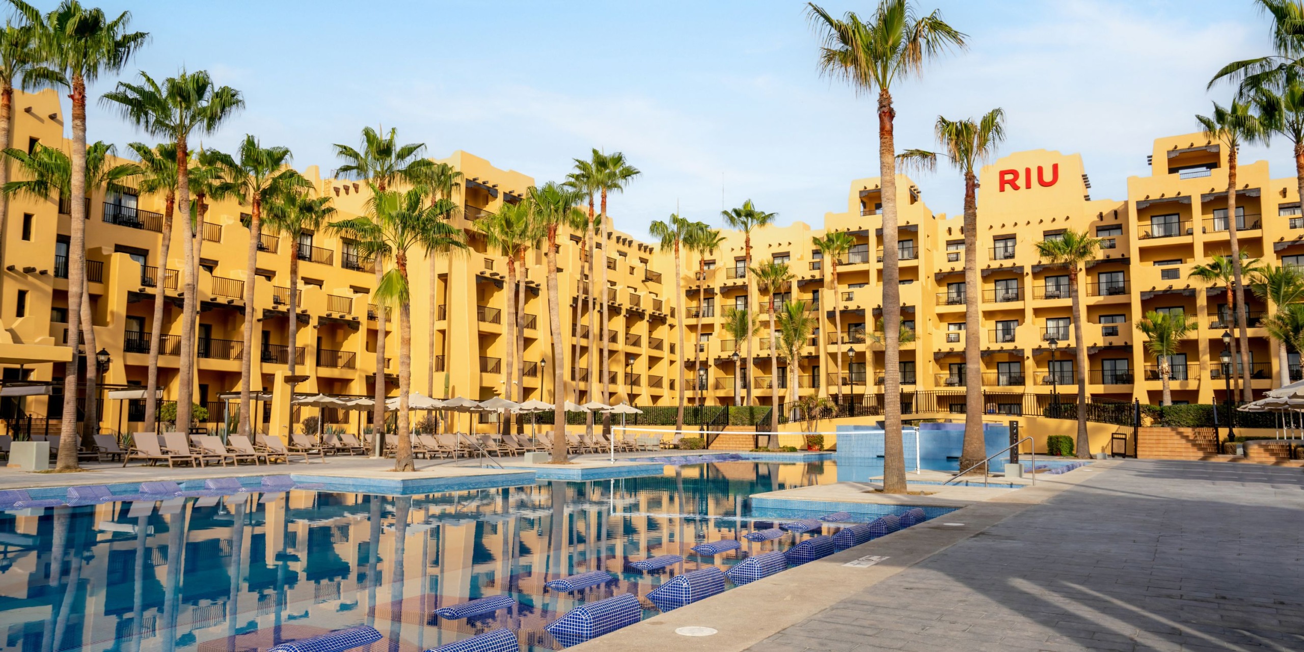 Hotel Riu Santa Fe, a spacious hotel featuring a yellow facade, surrounded by palm trees and boasting a swimming pool in front, all under a clear blue sky.