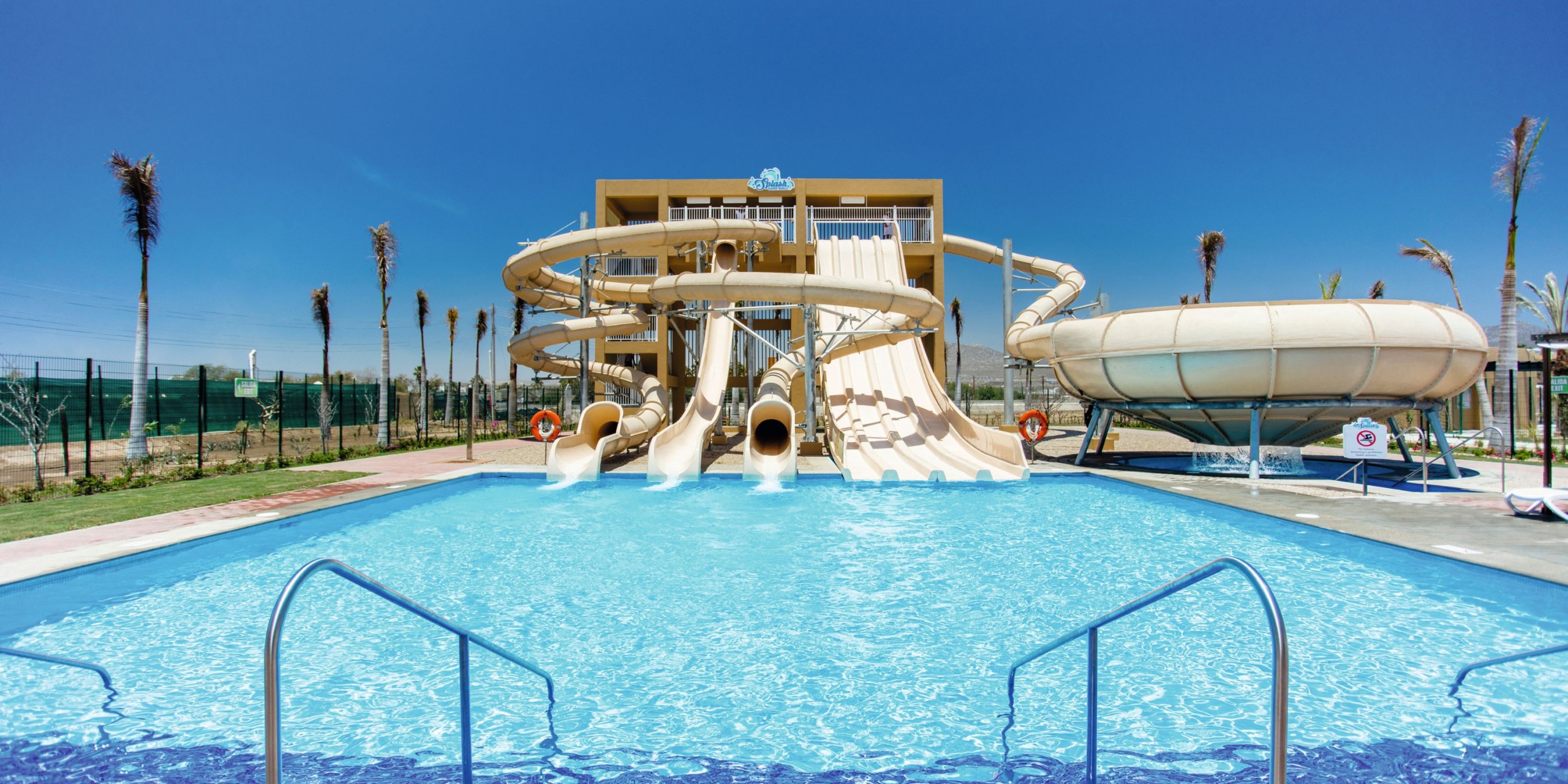 Experience the water park at Hotel Riu Santa Fe, where multiple curved slides lead into a large pool beneath a clear blue sky, with several palm trees lining the background.