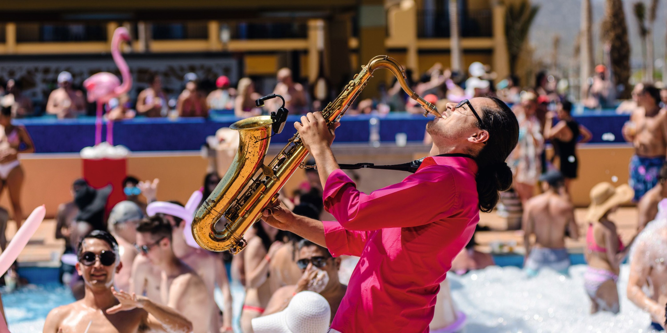 A musician in a pink shirt plays the saxophone by a crowded pool at Hotel Riu Santa Fe, where people in swimsuits are enjoying the sunny day and various pool activities in the background.