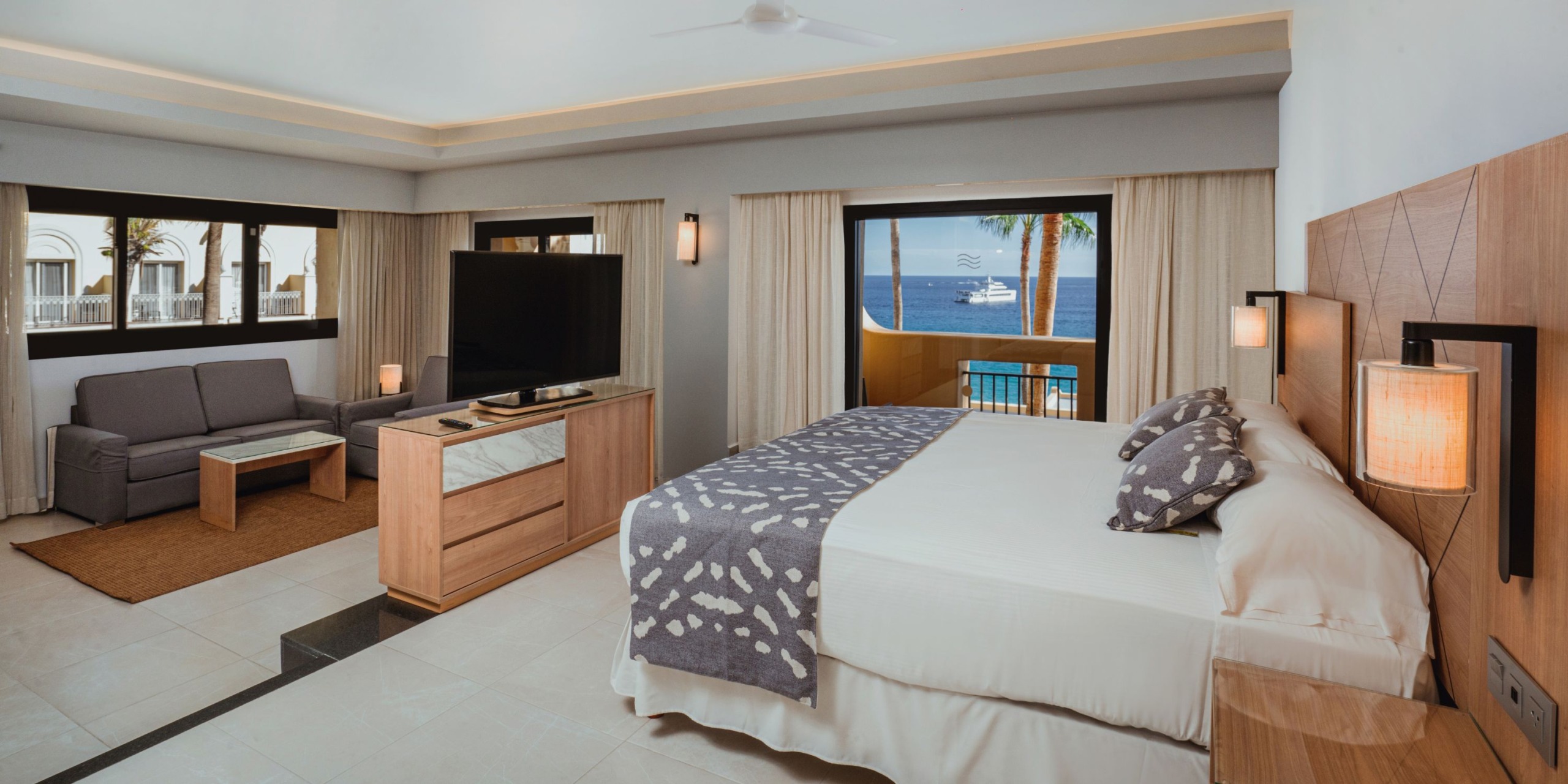 Enjoy the spacious accommodations at Hotel Riu Santa Fe, featuring a king-sized bed, a comfortable seating area, and a large television. Revel in the stunning ocean views through wide windows, all set against neutral tones and modern decor.