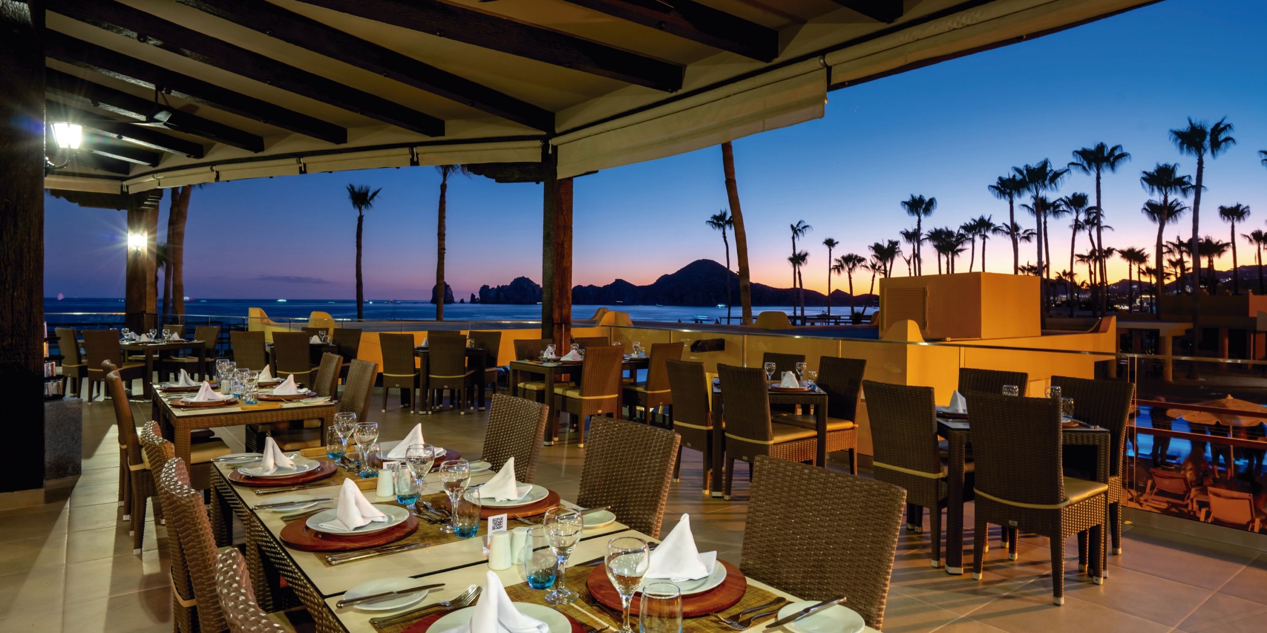 The terrace at Hotel Riu Santa Fe features set tables overlooking a pool and palm trees, all against the stunning backdrop of mountains and the ocean at sunset.