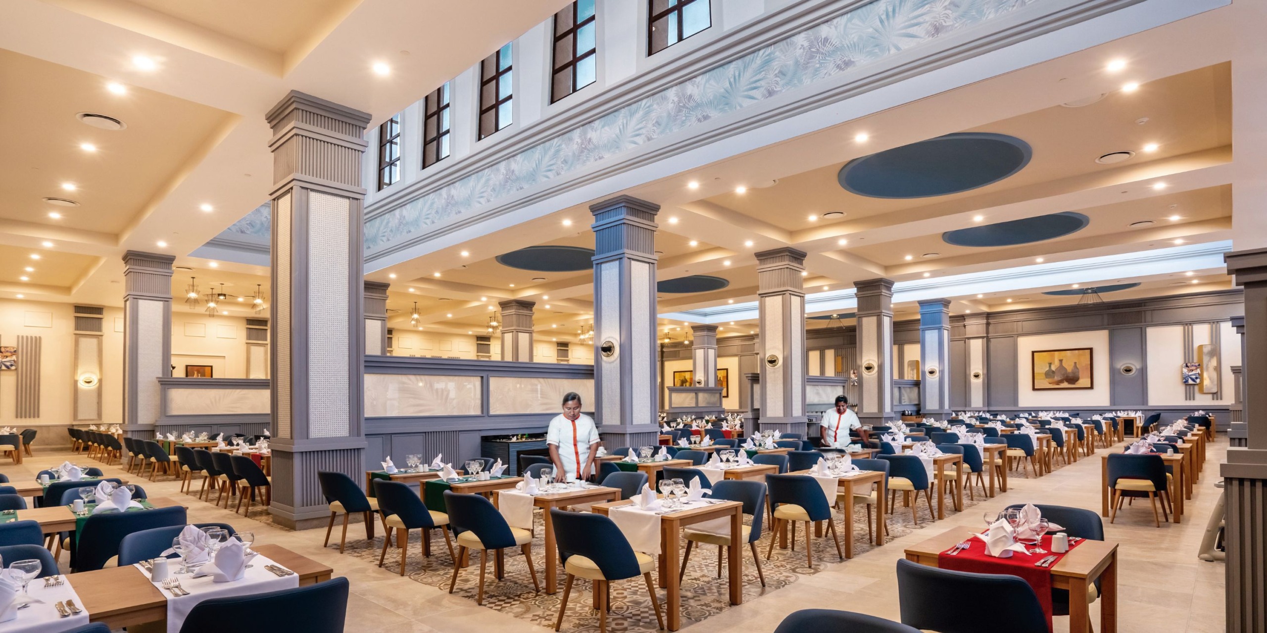 The interior of Hotel Riu Santa Fe boasts a spacious layout with high ceilings, elegant decor, and neatly arranged tables. Staff members in uniform diligently attend to the dining area.