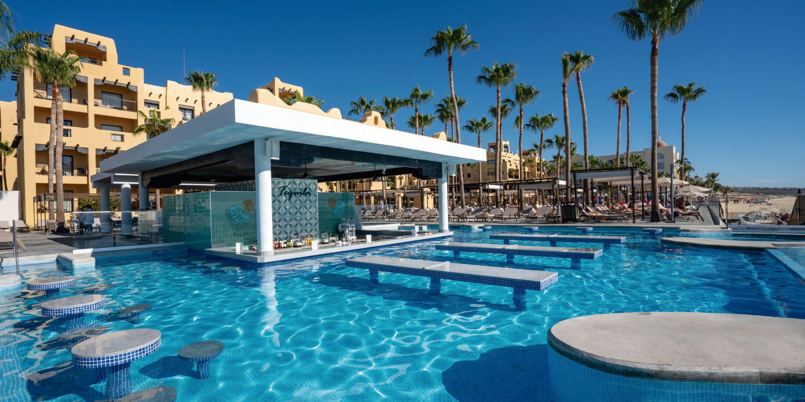 The Hotel Riu Santa Fe features a resort swimming pool complete with a central bar, bordered by comfortable lounge chairs and swaying palm trees, all beneath a clear blue sky.