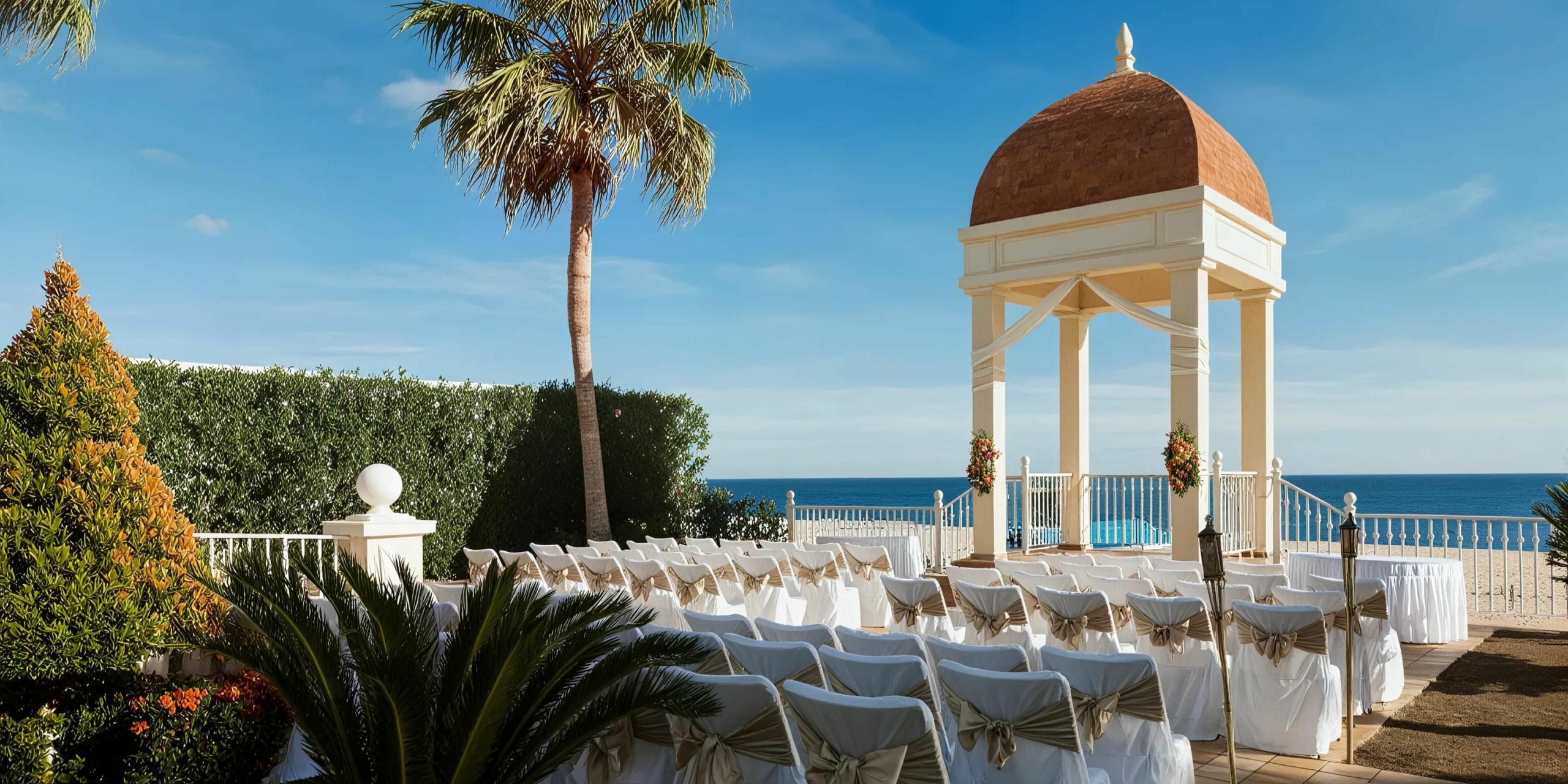 An outdoor wedding setup at Hotel Riu Palace Cabo San Lucas features rows of white chairs facing a gazebo by the sea, adorned with beige ribbons and floral decorations. Tall palm trees and a clear blue sky create a stunning backdrop.