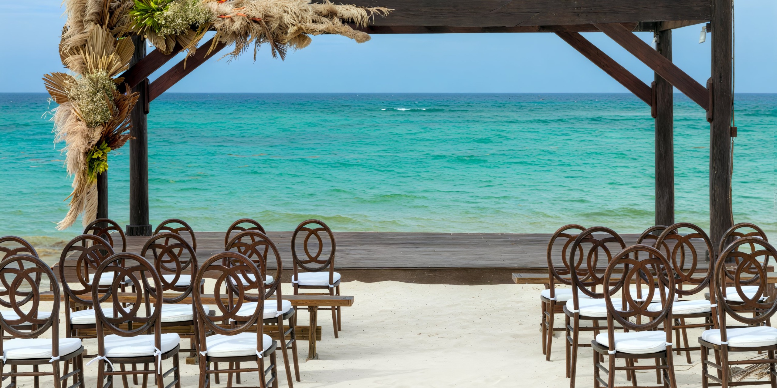 A destination wedding at Royalton Bavaro features a stunning setup with wooden chairs and an elegant arch, all with a breathtaking view of the turquoise ocean.