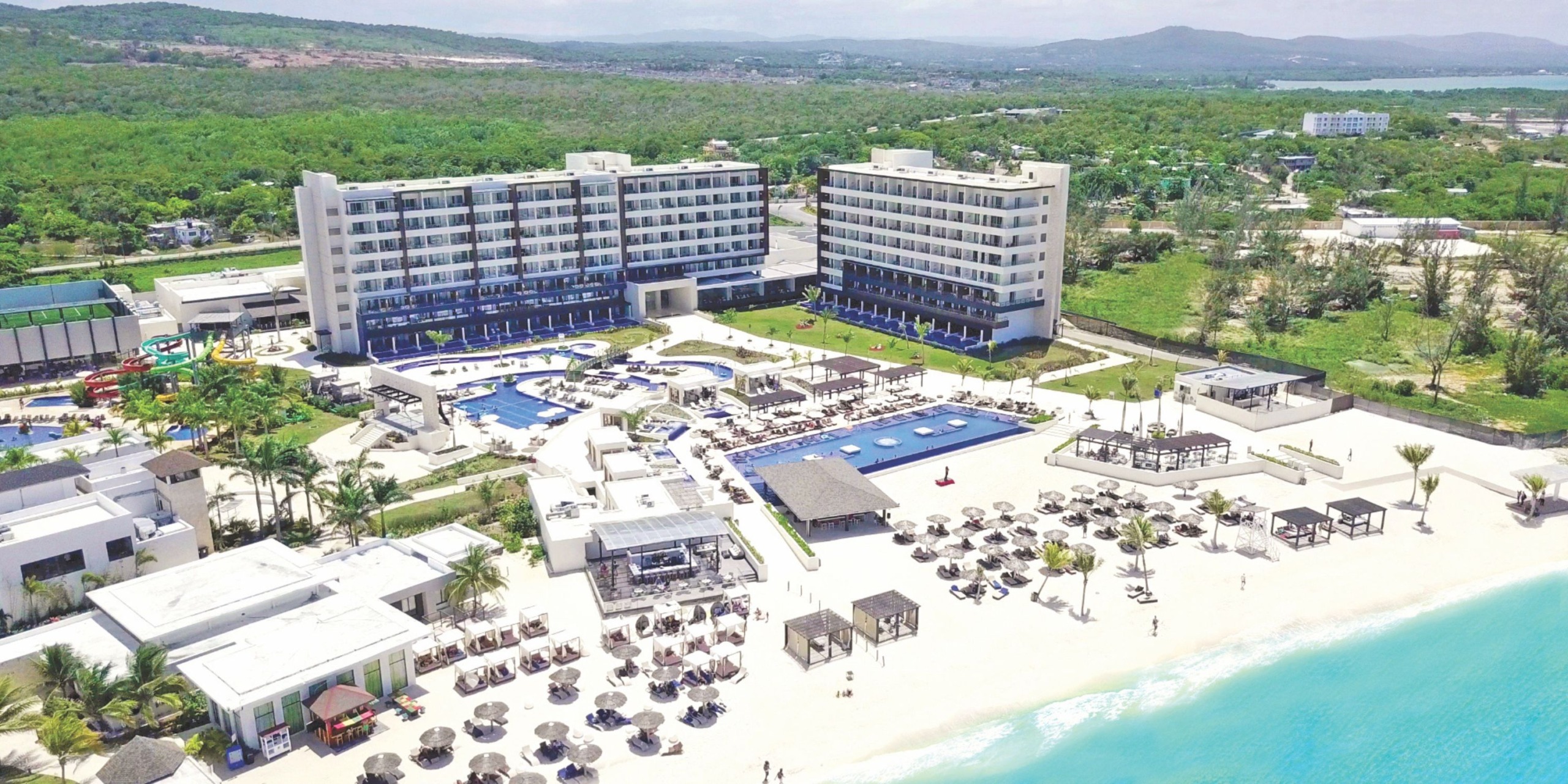 Aerial view of the Royalton Blue Waters, a beachfront resort featuring two large buildings, pools, sun loungers, and palm trees. The ocean is visible on the right, with green hills providing a picturesque backdrop in the distance.