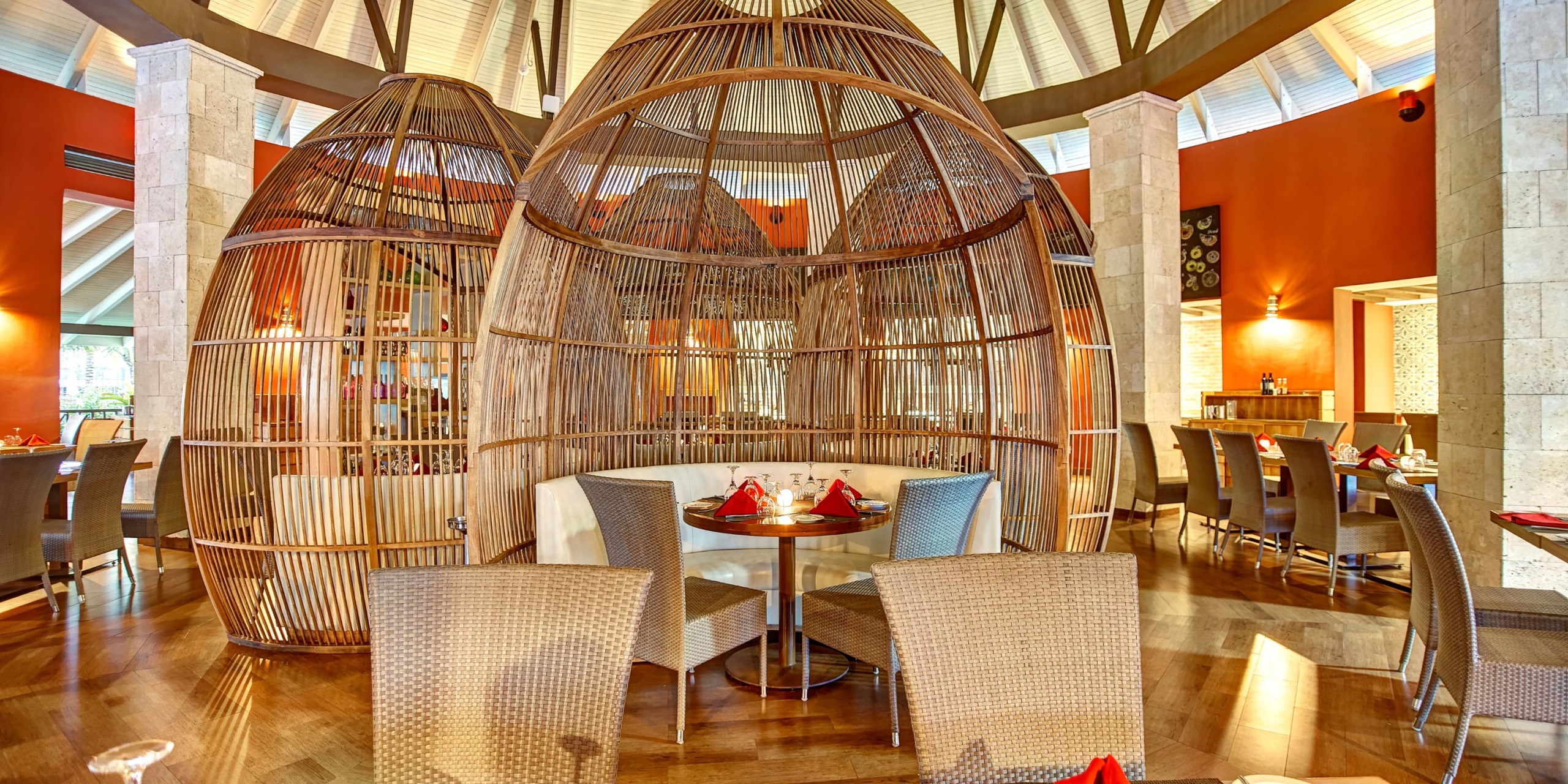 The elegant interior of the Royalton Blue Waters features circular bamboo seating pods, a wooden floor, and orange walls. The tables are adorned with red napkins and glasses.