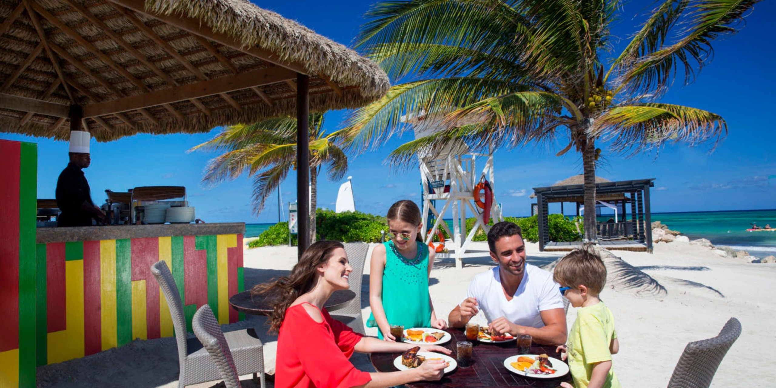 A family of four enjoys a meal at the Royalton Blue Waters restaurant, surrounded by palm trees and with the ocean in the background.