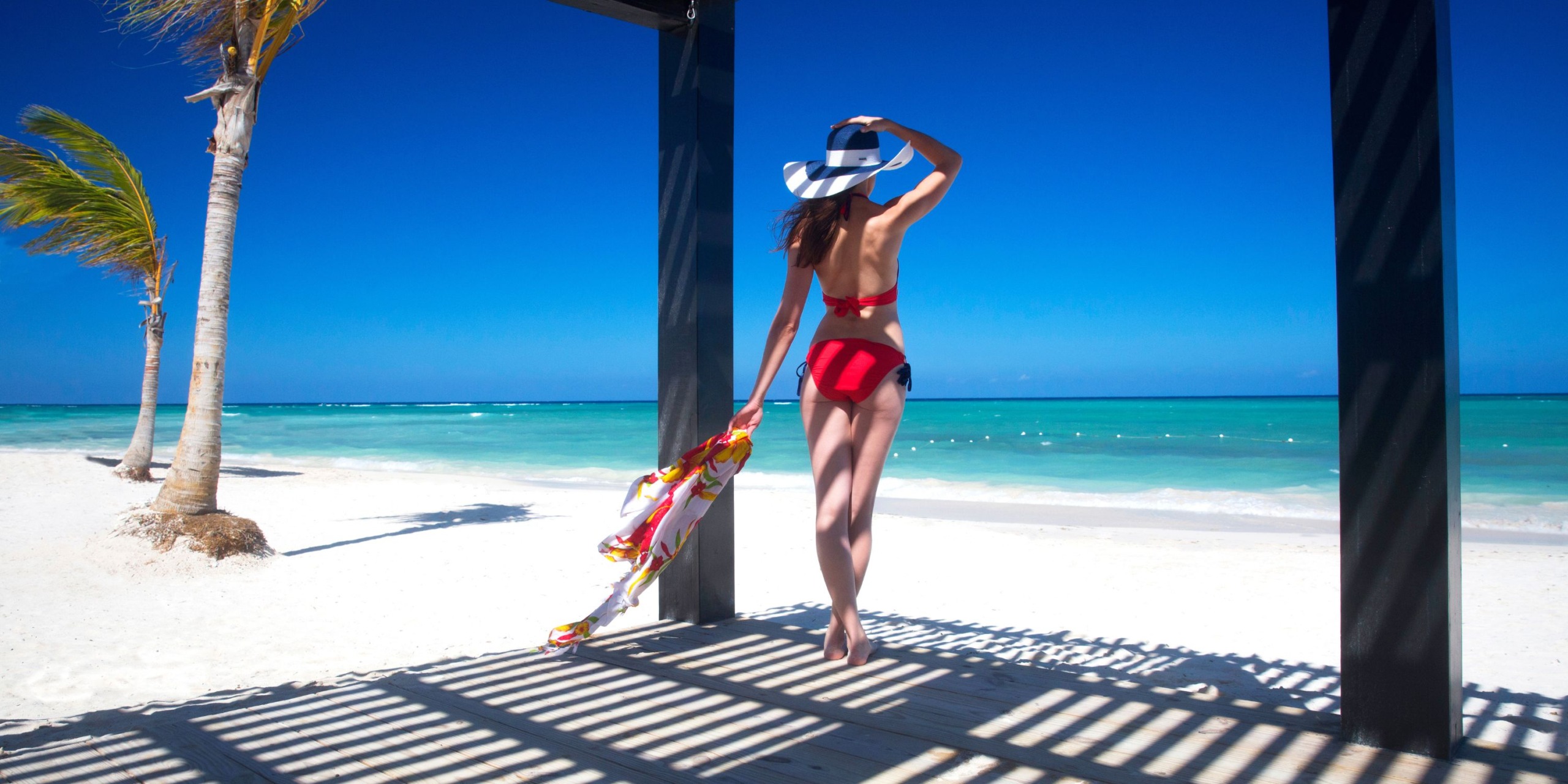 A woman in a red bikini and hat stands under a pergola at Royalton Blue Waters, gazing at the turquoise ocean on a sunny beach, while holding a colorful sarong.