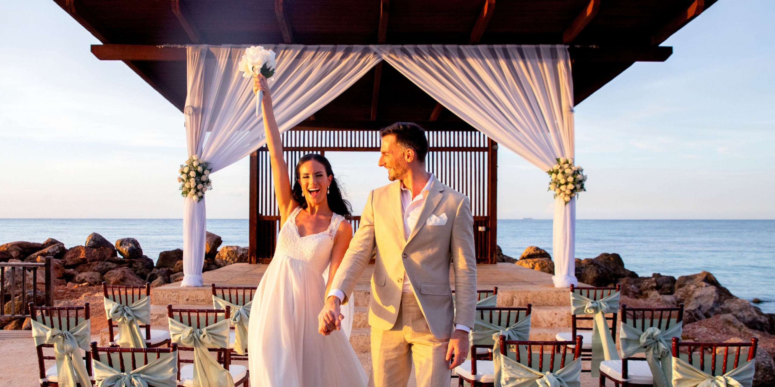 A couple celebrates their wedding in front of a decorated gazebo by the ocean at Royalton Blue Waters. She raises her bouquet in joy.