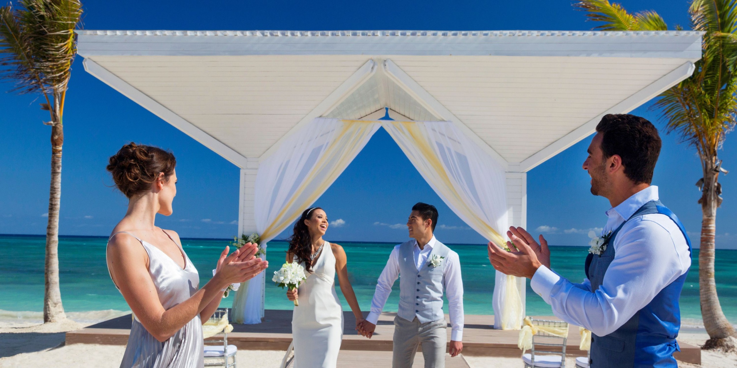 A newly married couple stands hand in hand under a beachside gazebo at Royalton Blue Waters, surrounded by two applauding guests, with palm trees and the ocean in the background.