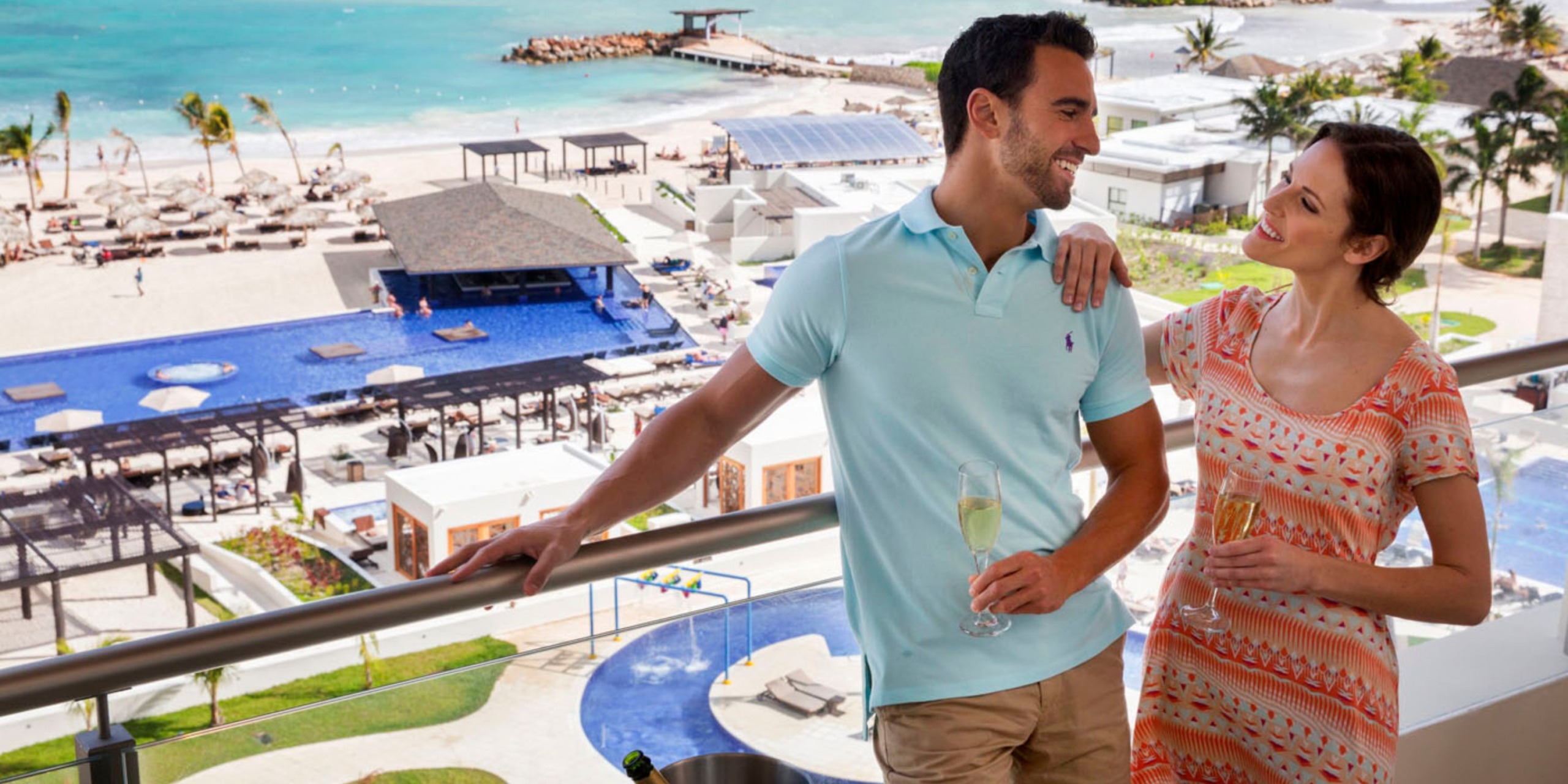A couple stands on a balcony at the Royalton Blue Waters, overlooking a beach resort with a pool and lounge chairs, holding champagne glasses.